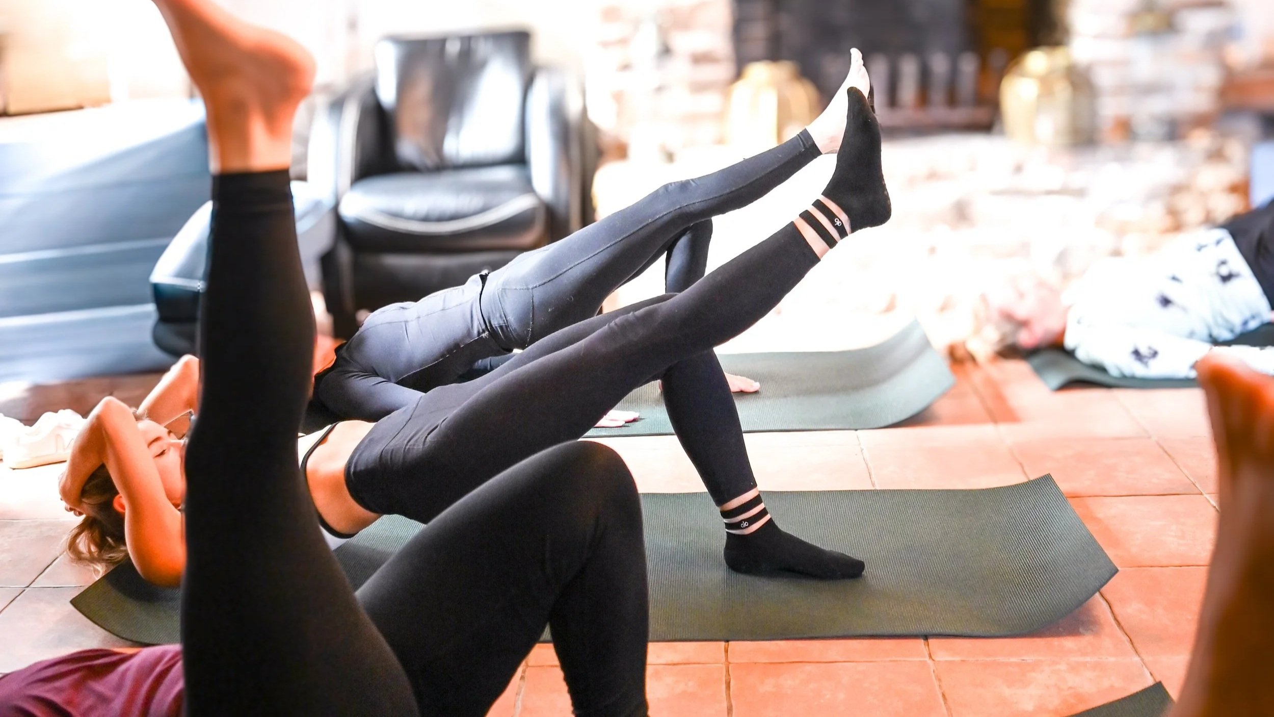 Women participating in a yoga class, lying on yoga mats on a tiled floor, in a cozy indoor setting.