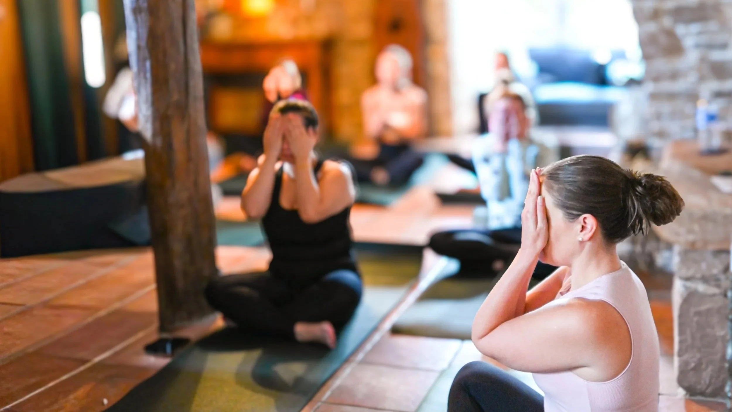 Group of women practicing yoga or meditation in a cozy, rustic indoor setting, sitting cross-legged on yoga mats with their hands covering their faces in a meditative pose.