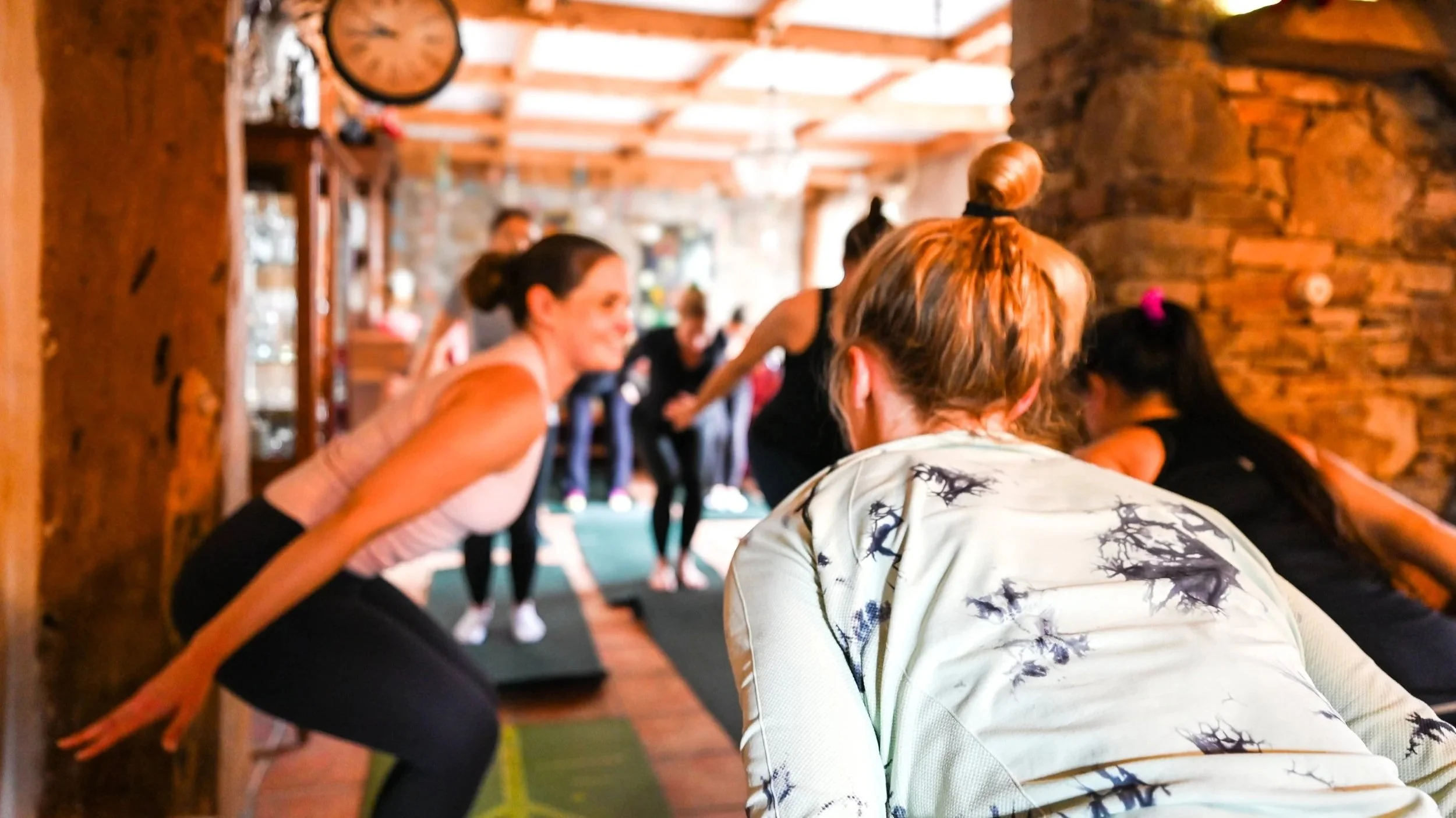 Group of women in a yoga class performing stretches in a rustic indoor setting with wooden walls and a stone fireplace.