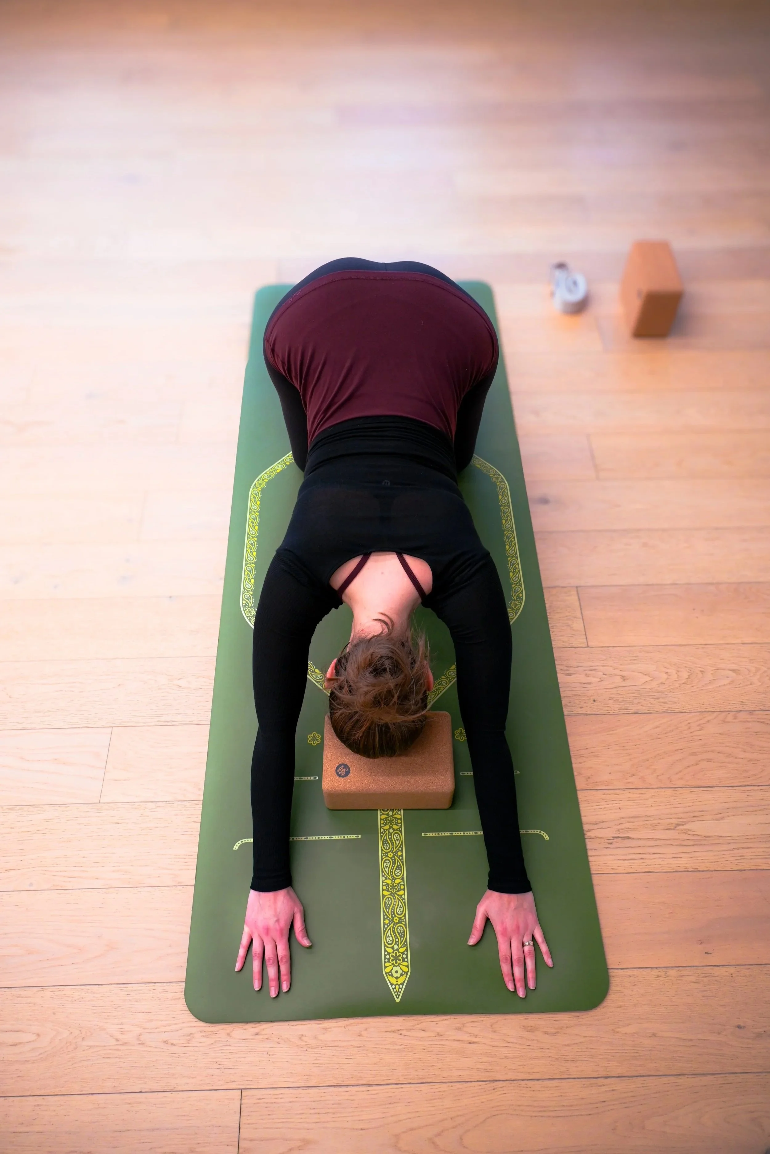 A woman practicing yoga on a green mat, bending forward with her head and arms resting on a cork block, in a room with wooden flooring.
