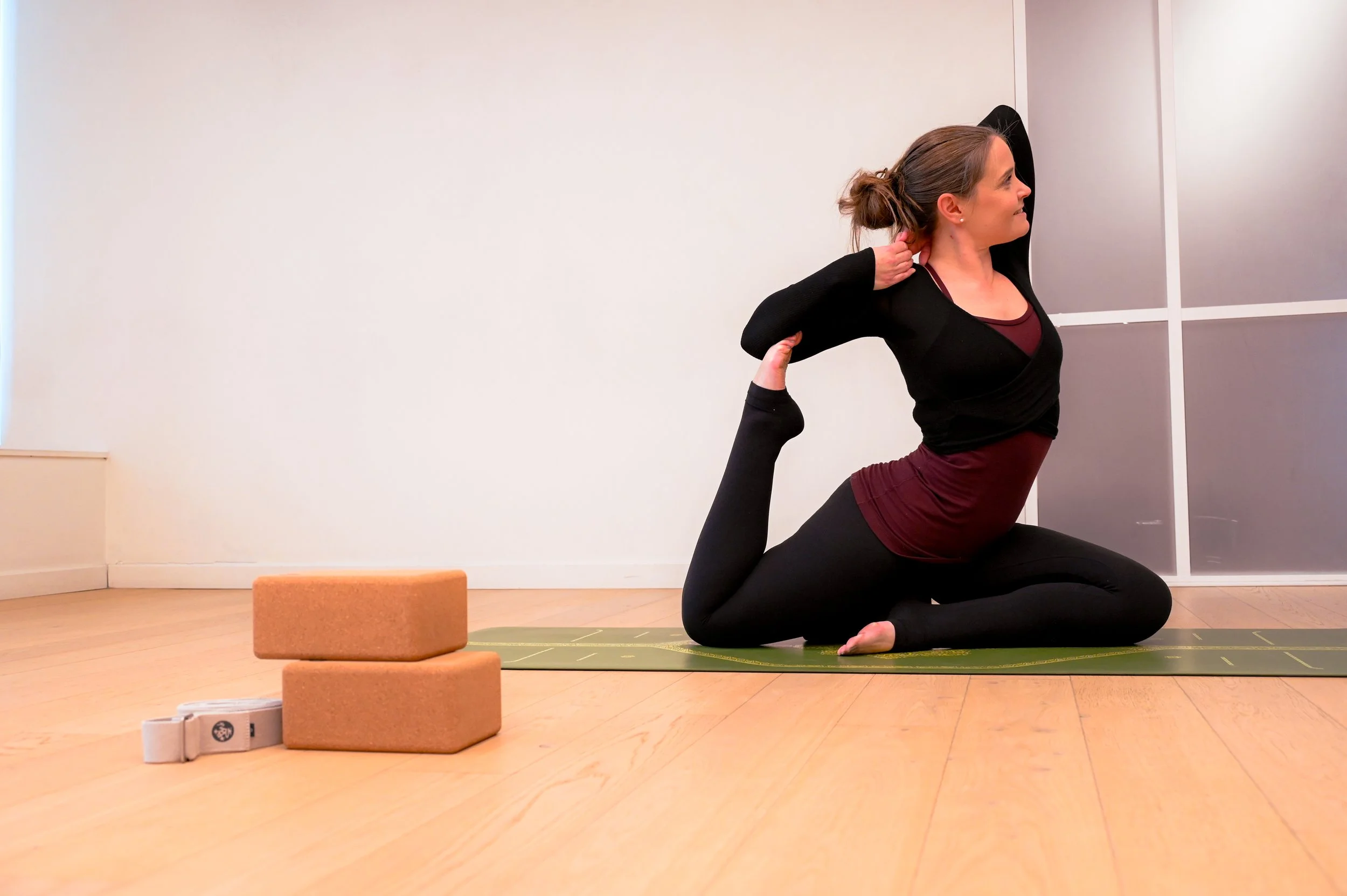 A woman practicing yoga indoors, performing a pose on a green yoga mat, with two cork yoga blocks nearby.