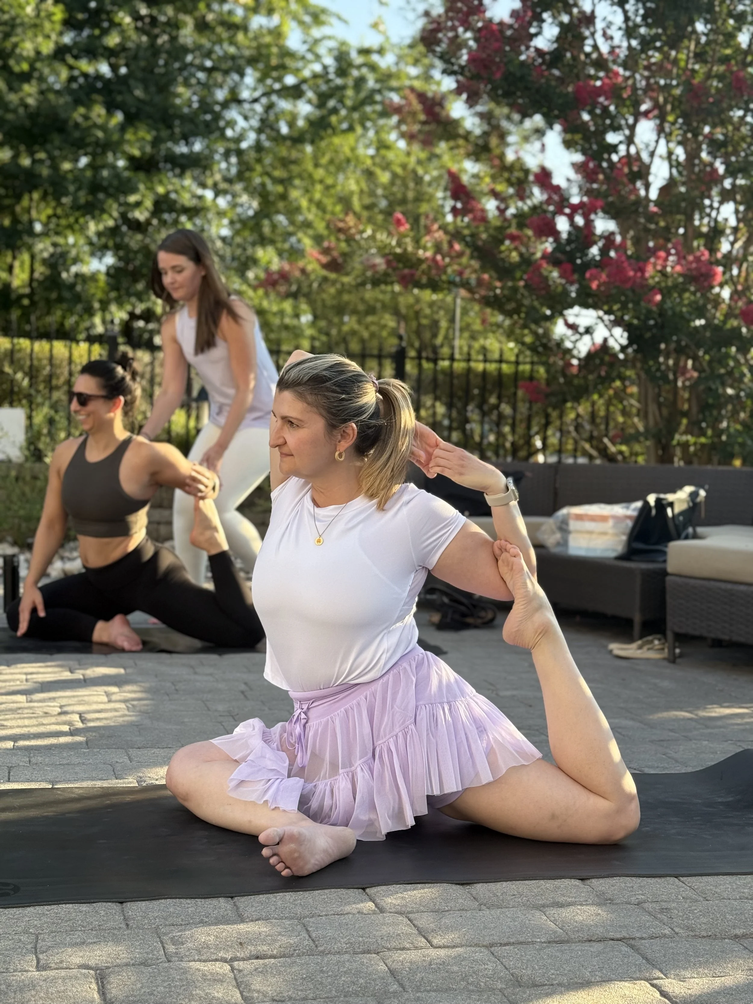 Four women practicing yoga outdoors on a sunny day, with trees and pink flowers in the background. One woman in the foreground is in a seated spinal twist pose on a yoga mat, wearing a white top and a lavender skirt. Two women in the background are assisting and another is kneeling in a yoga pose.