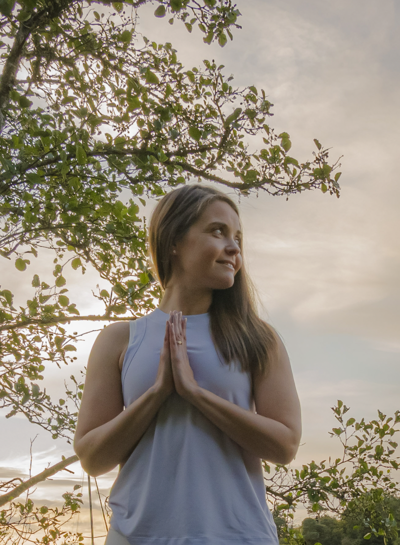 A woman practicing yoga outdoors during sunset, standing with hands in prayer position near her chest, surrounded by trees and a peaceful sky.