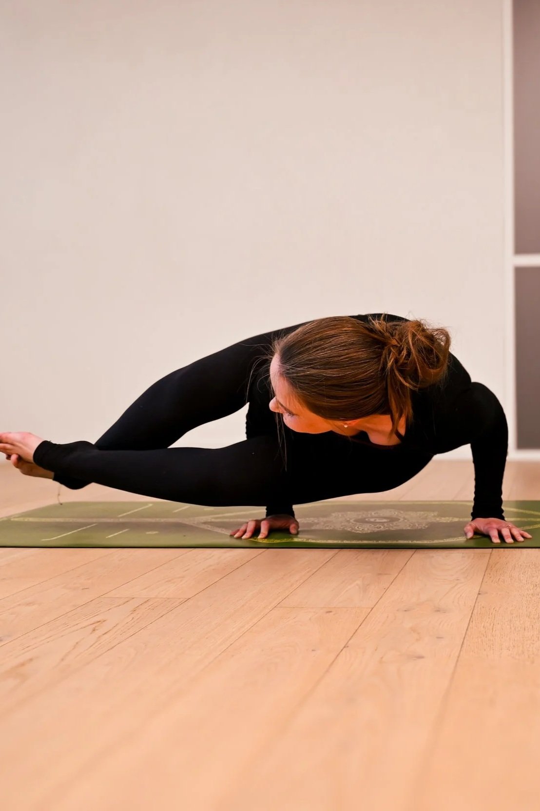 A woman practicing yoga on a mat indoors with a white wall background, performing a challenging arm balancing pose (8 angle pose)