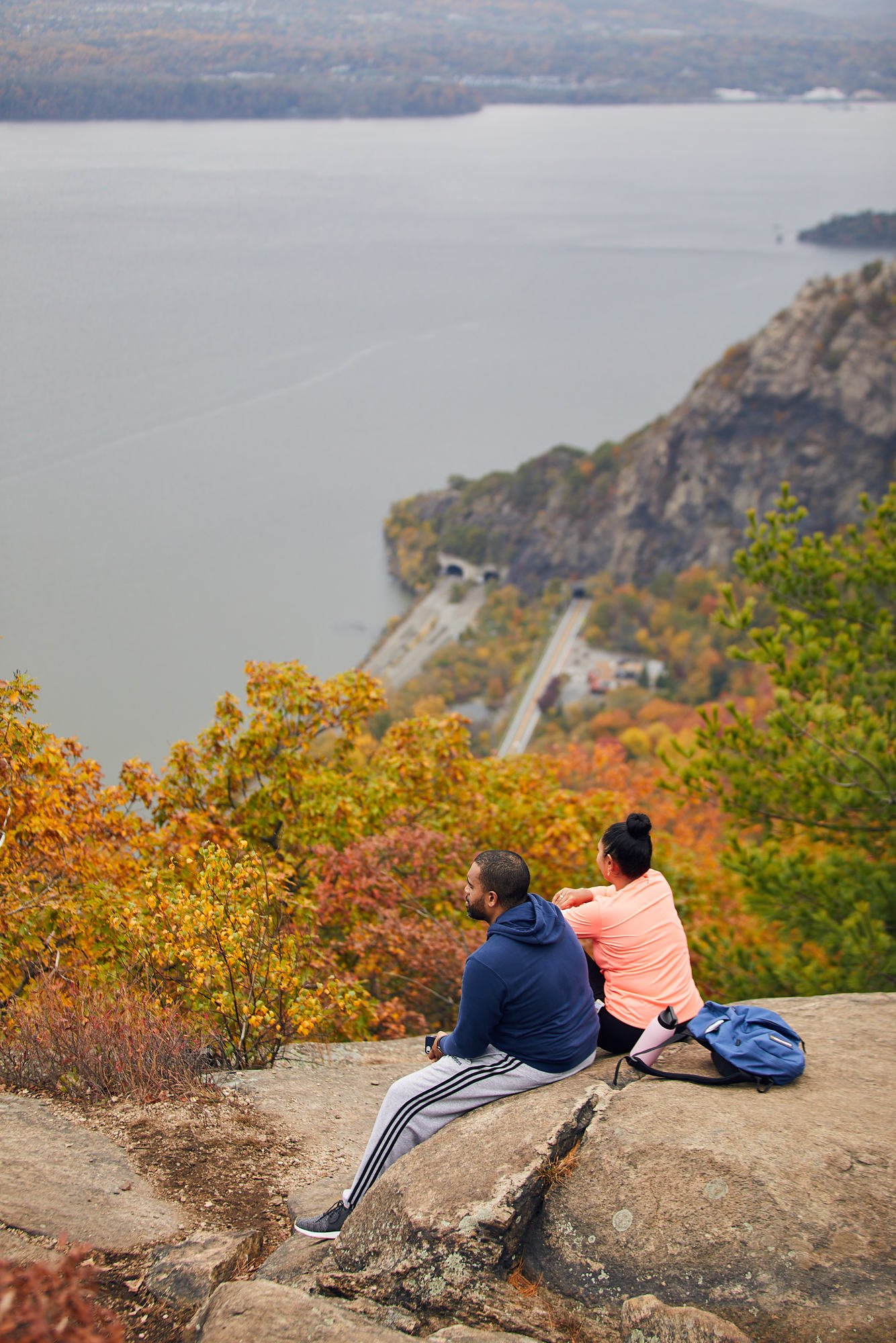 Two people sitting on a rock at the top of Bull Hill overlooking a view of an autumnal landscape with a river, Breakneck Ridge first false summit., trees with fall colors, and Route 9D going through Breakneck tunnel below