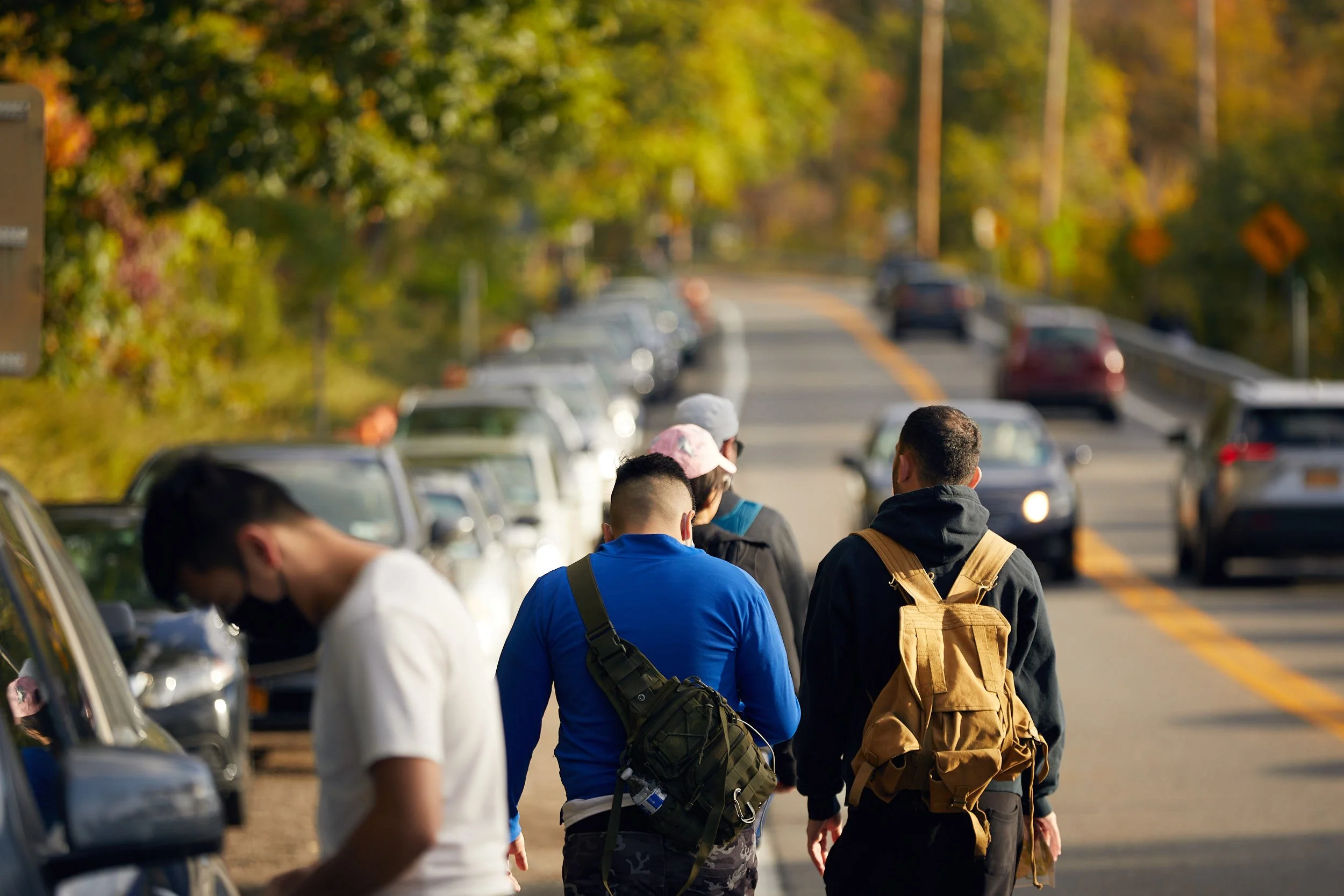People walking to Breakneck Ridge on busy Route 9D with parked cars, passing cars, and greenery in the background.
