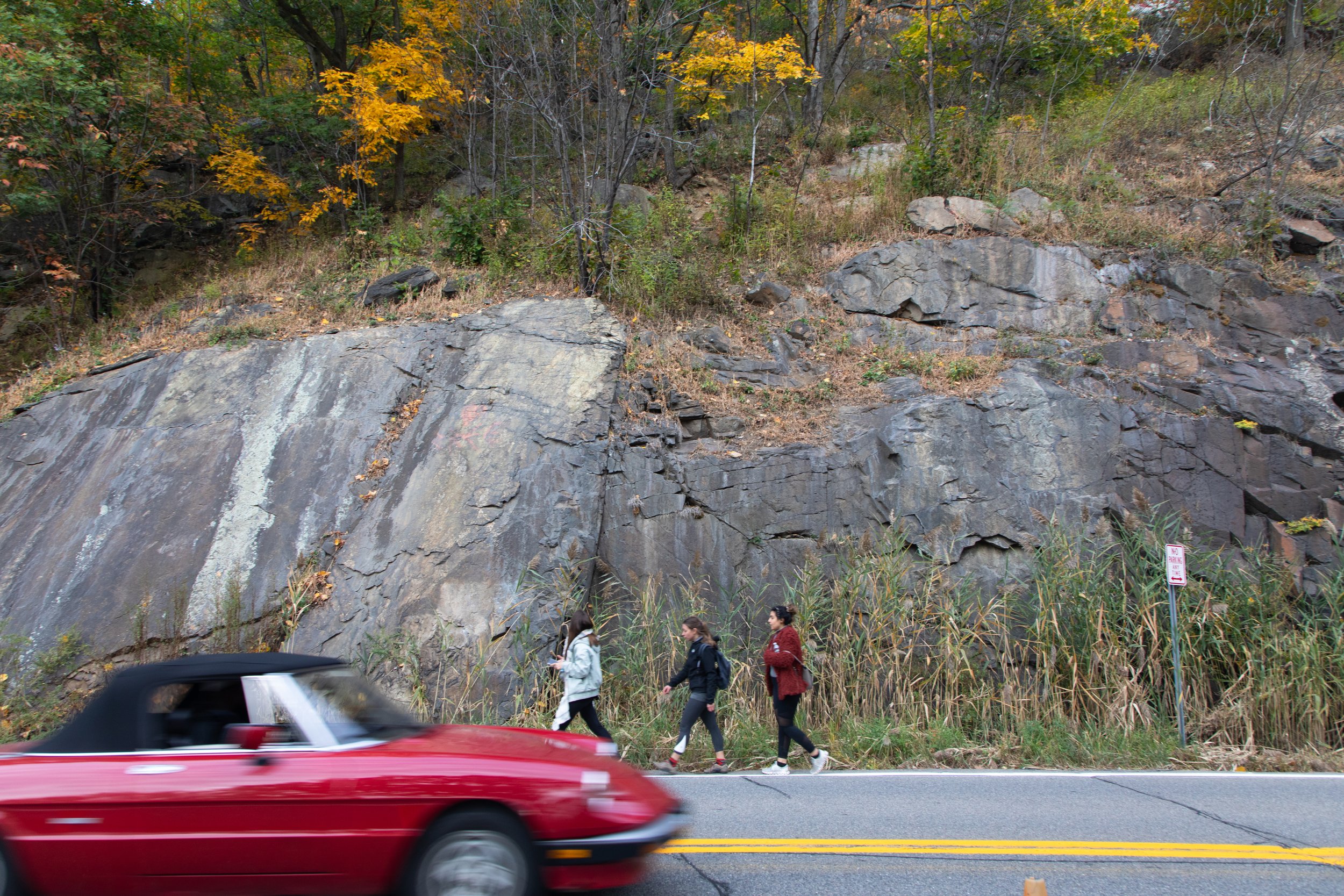 Three pedestrians walking alongside the rocky roadside of Route 9D with autumn foliage, as a red convertible car passes by on the road going south toward Breakneck Ridge.