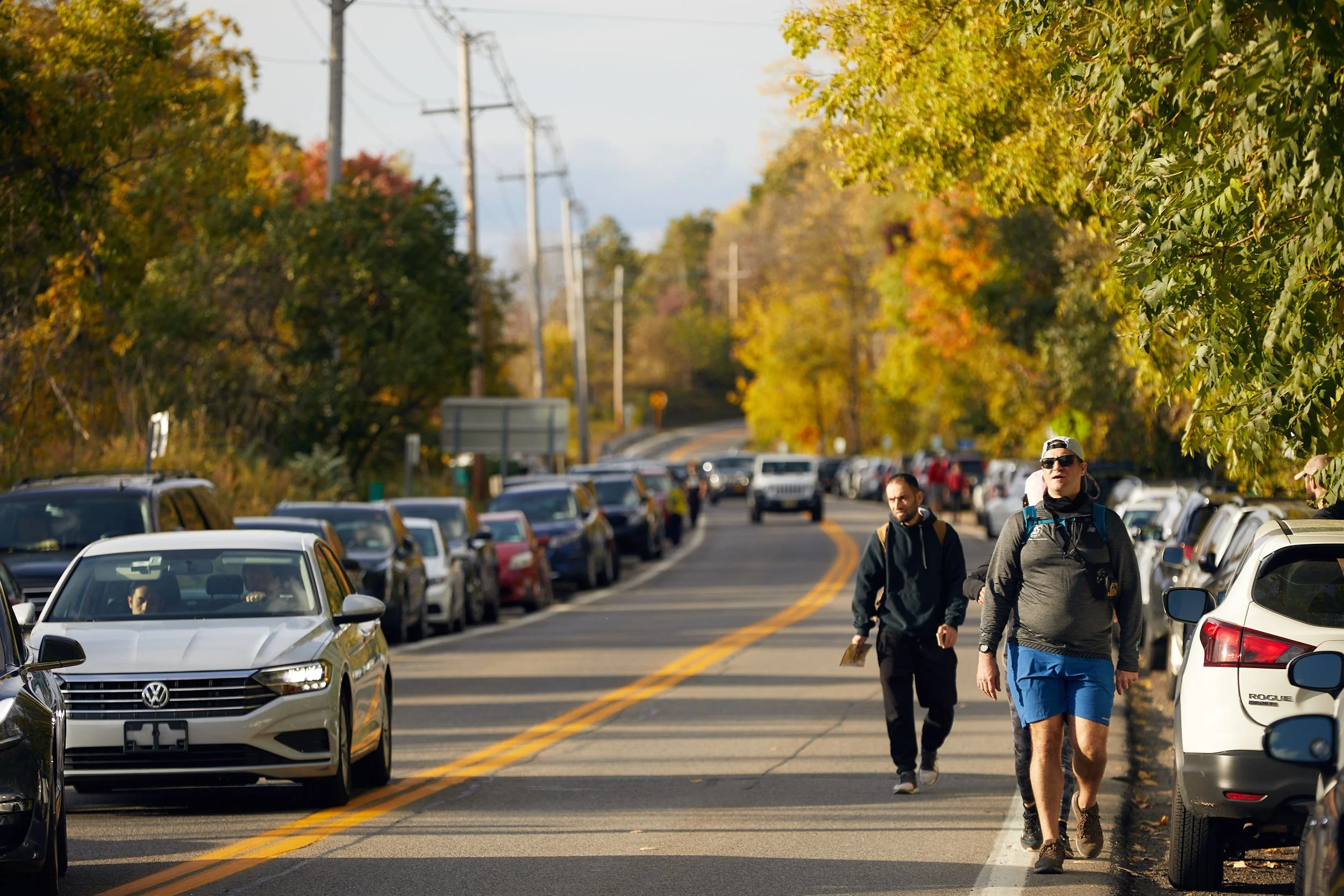 People walking in the road (Route 9D) toward Breakneck Ridge. Road is lined with parked cars and surrounded by autumn trees.