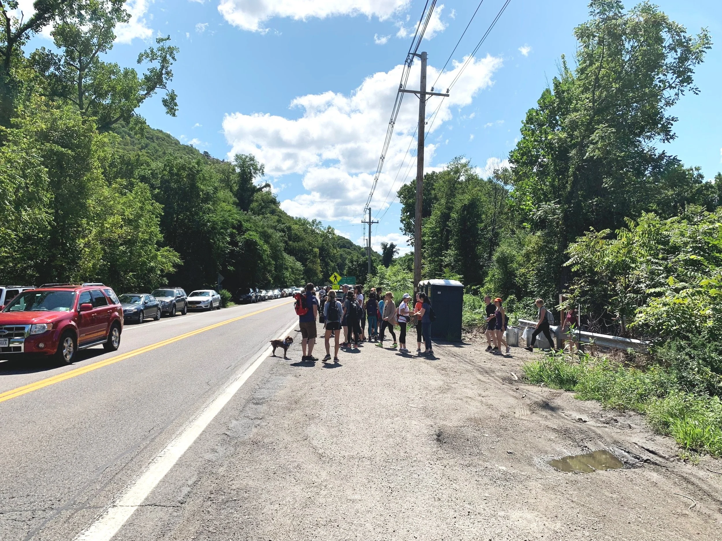 Group of people with backpacks and a dog standing near the roadside of Route 9D, with parked cars, cars passing by, a port-a-potty, and surrounded by trees and a blue sky with clouds. Hikers preparing to hike at Breakneck Ridge and Wilkinson Memorial