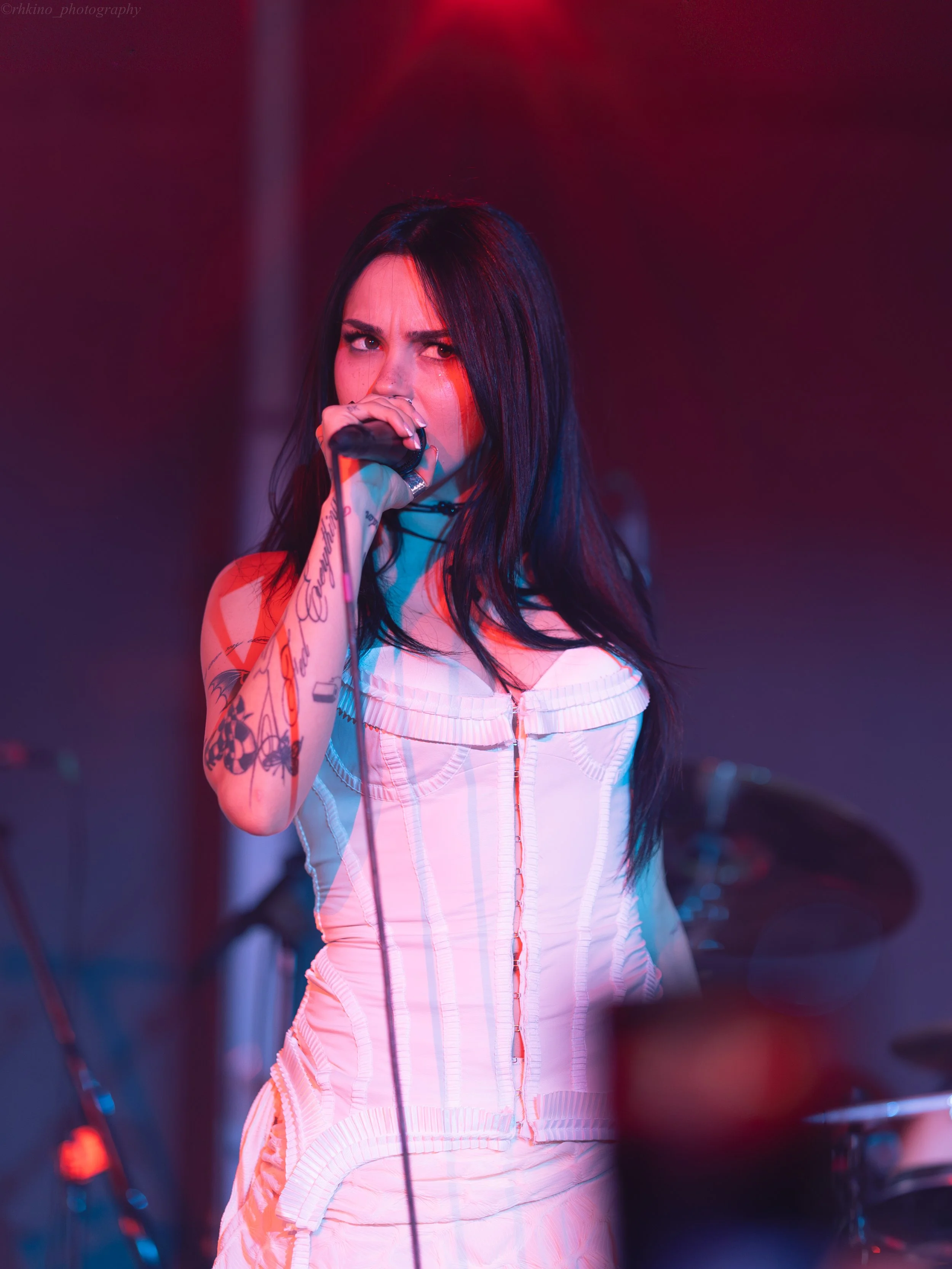 A woman with long dark hair and tattoos on her arm is singing into a microphone on stage, with colorful pink and blue lighting.