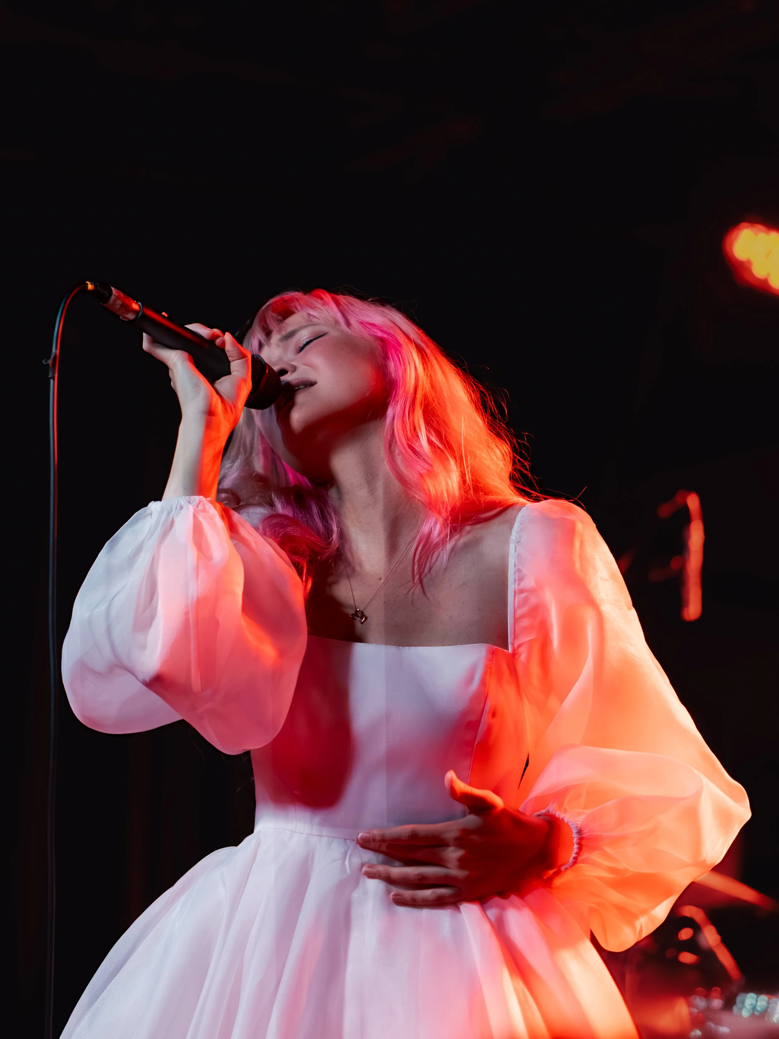 A woman with pink hair singing into a microphone on stage, wearing a white dress with puffy sleeves.