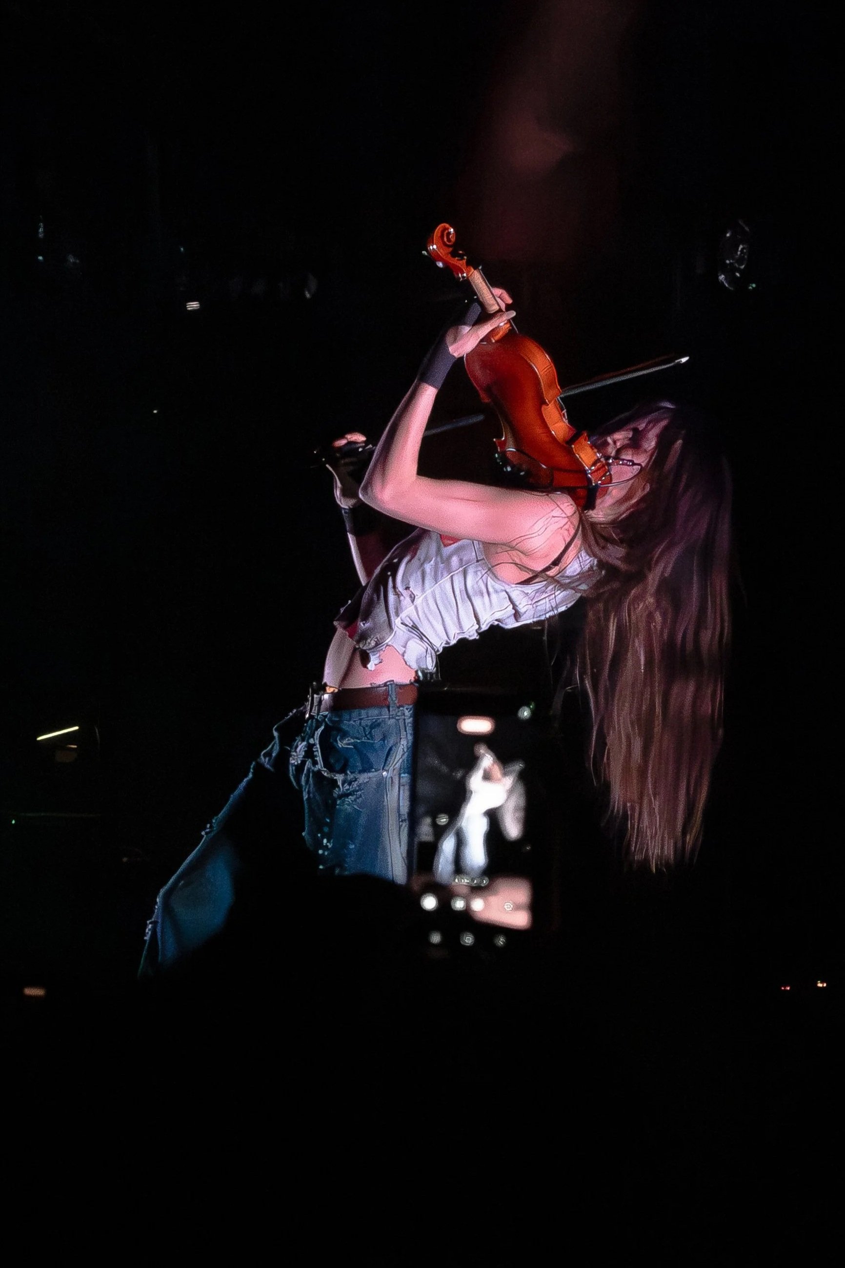 A woman with long hair performing on stage, holding a violin and bow, dressed in a sleeveless top and ripped jeans, with a dark background.
