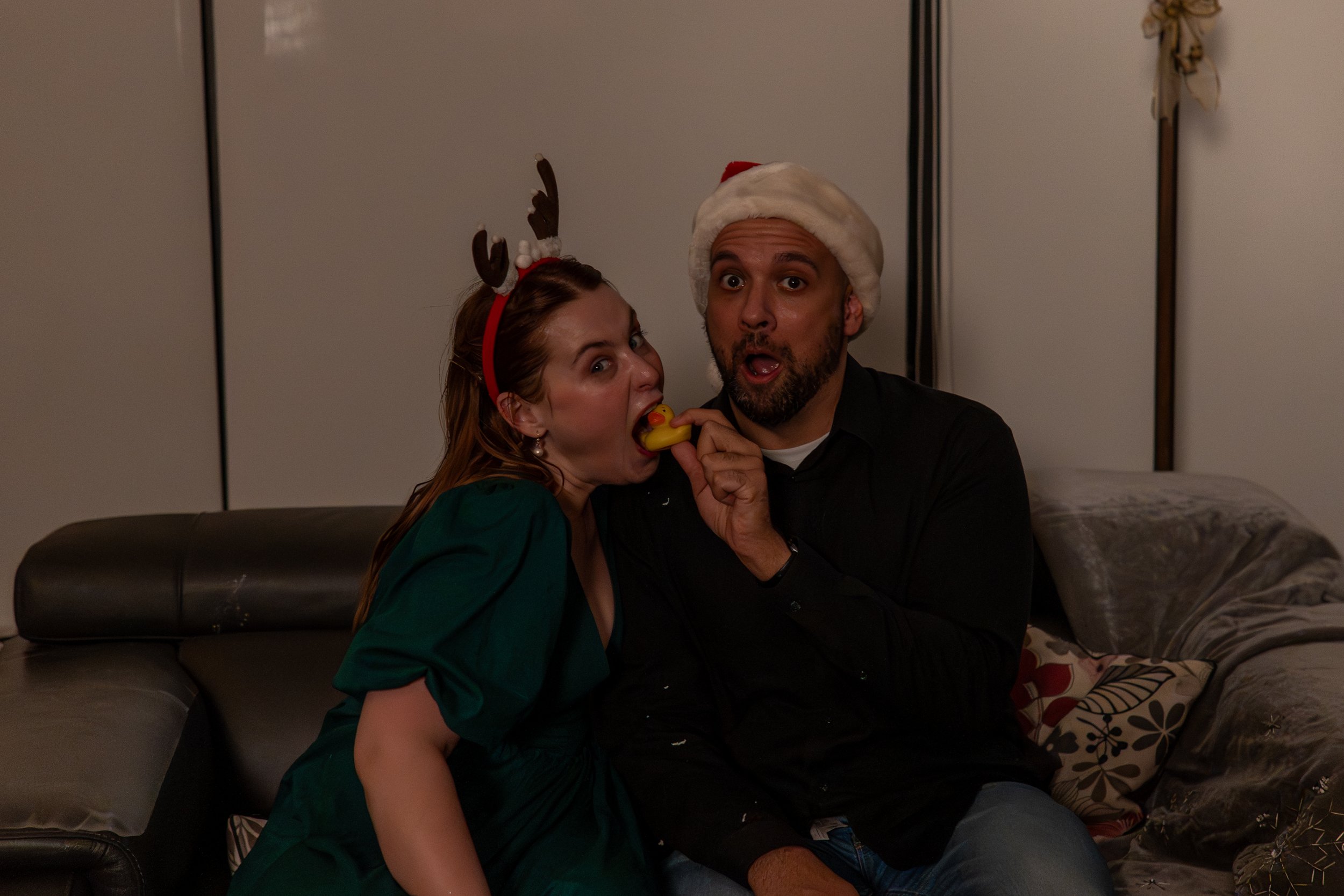 A woman and a man wearing a Santa hat and reindeer antlers are sitting together on a couch, with the woman licking a rubber duck while the man holds it. They are in a cozy indoor setting with pillows and a decorated wall.