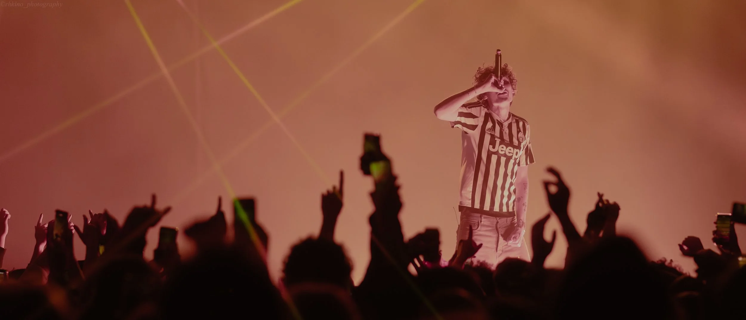 A musician performs on stage with orange lighting and fog; he is wearing a striped Juventus football jersey and is holding a microphone to his mouth, with a crowd in front raising their hands and phones.