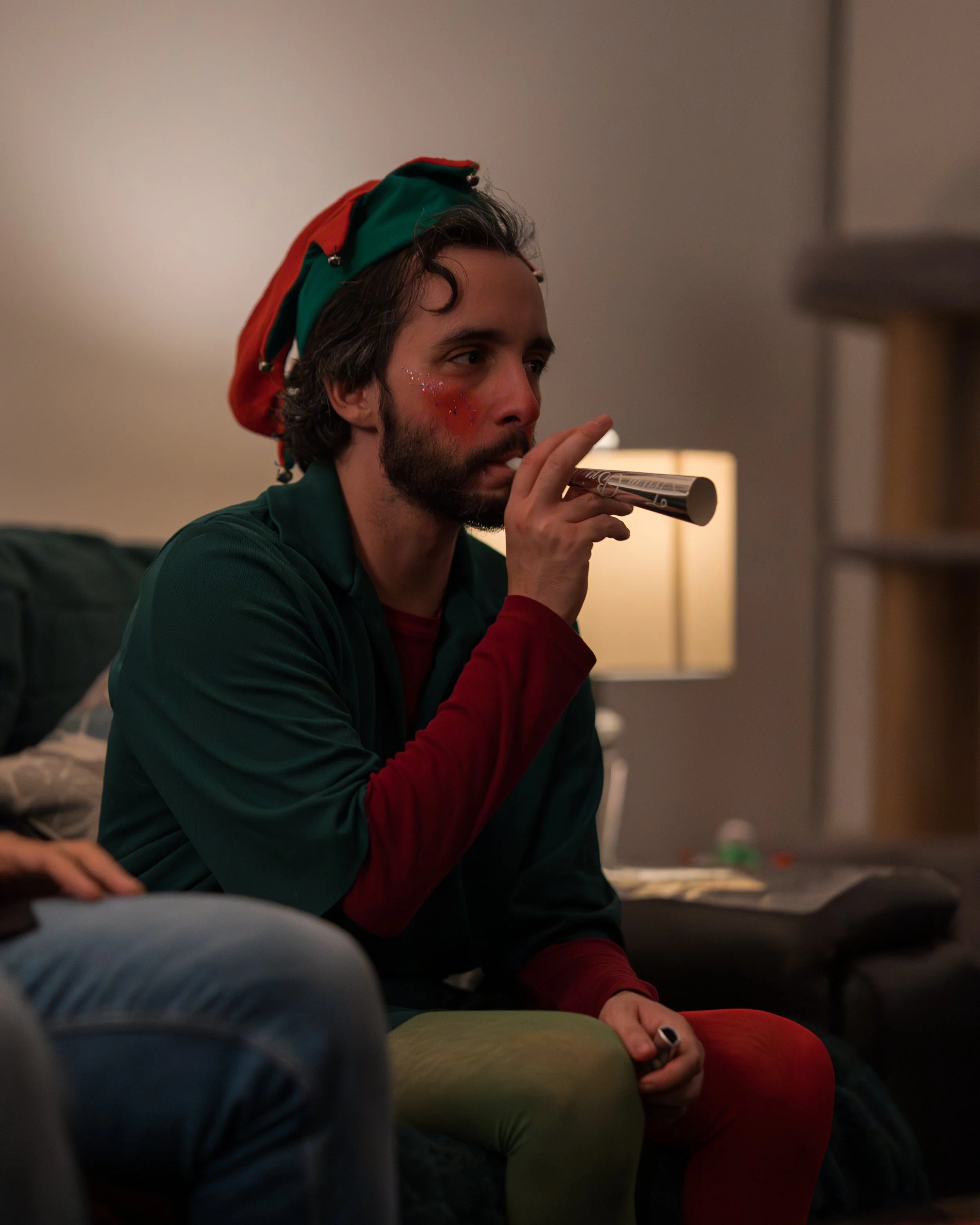 A man with curly hair and a beard wearing a festive red and green elf hat, and red glitter makeup on his cheek, is sitting on a couch blowing into a metallic party horn in a dimly lit room.