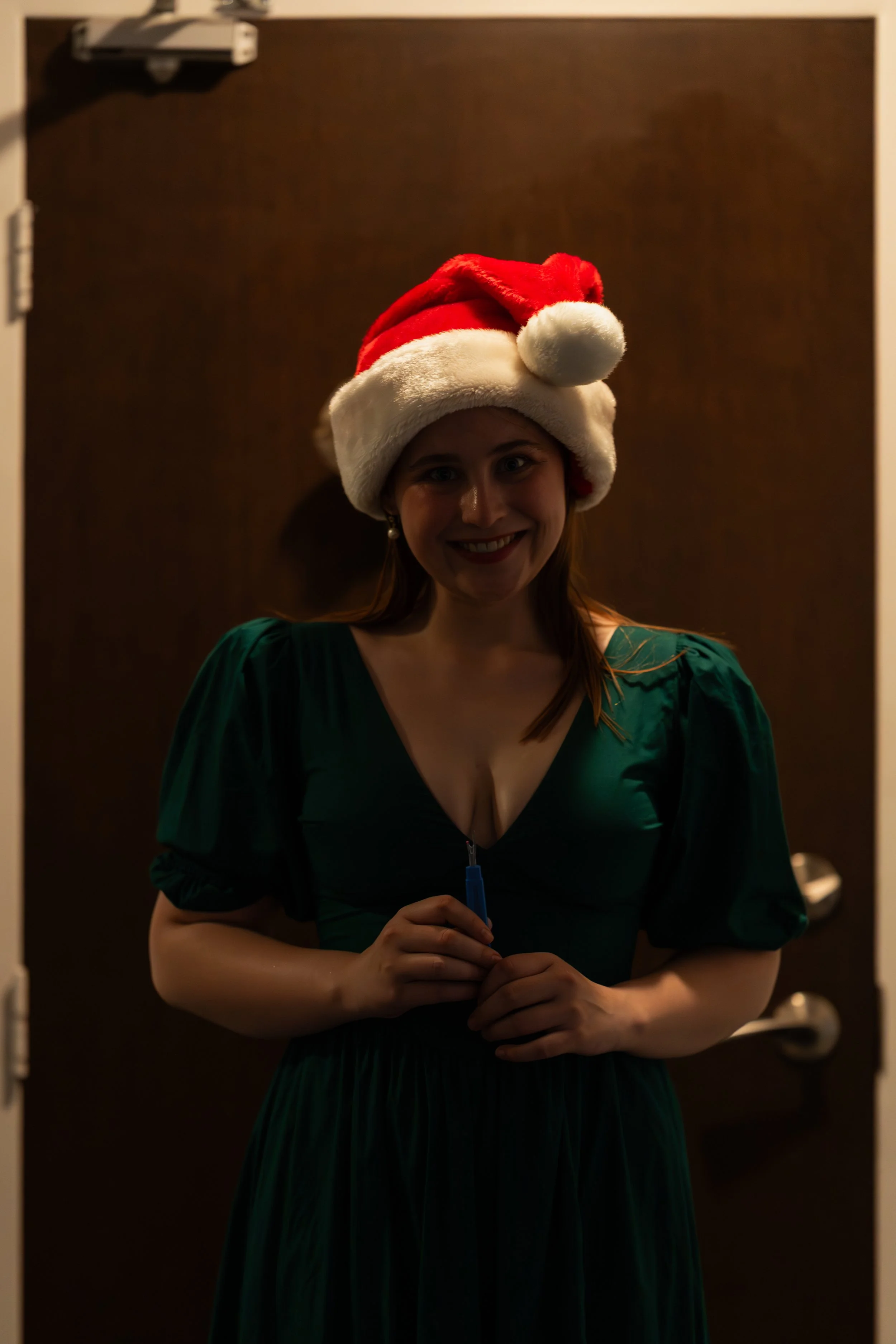 A woman wearing a Santa hat and a green dress, smiling and holding a pen, standing in front of a dark brown door.