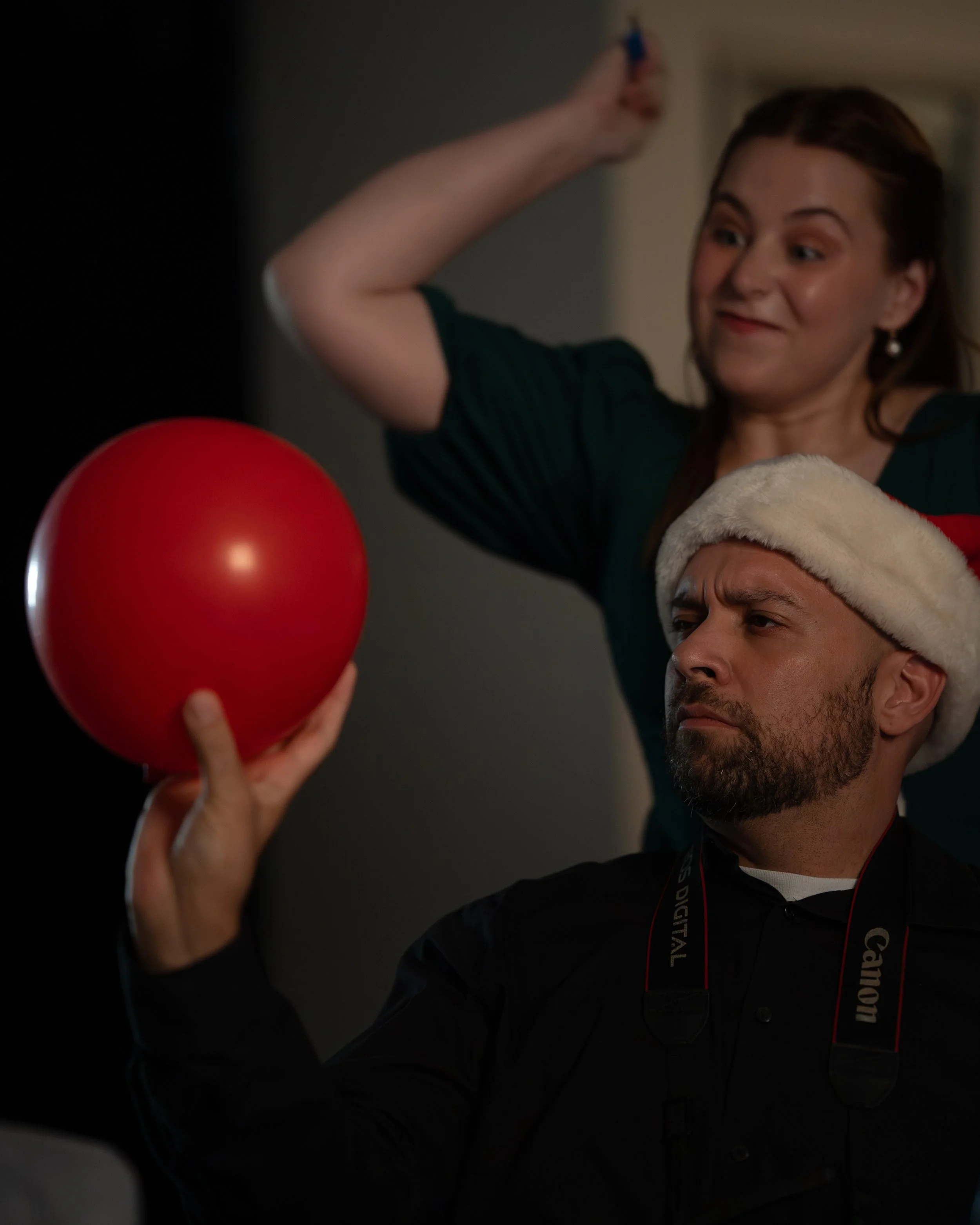 A woman with a festive shirt and earrings stands behind a man wearing a Santa hat, who is holding a red ball and looking at it with a focused expression.