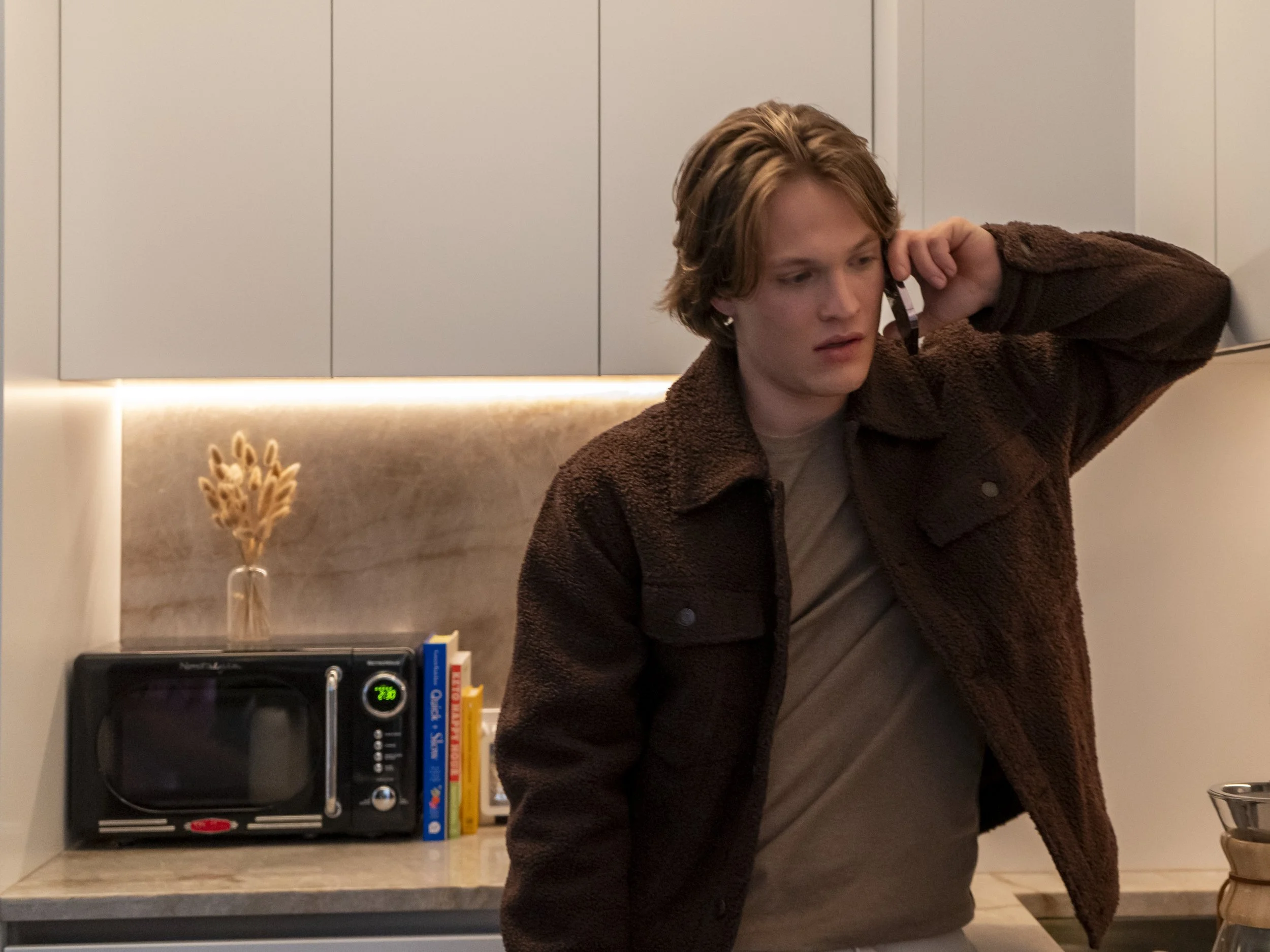 A young man with red hair talking on the phone in a modern kitchen. He is wearing a brown jacket and a beige shirt, standing near a counter with books and a microwave oven.