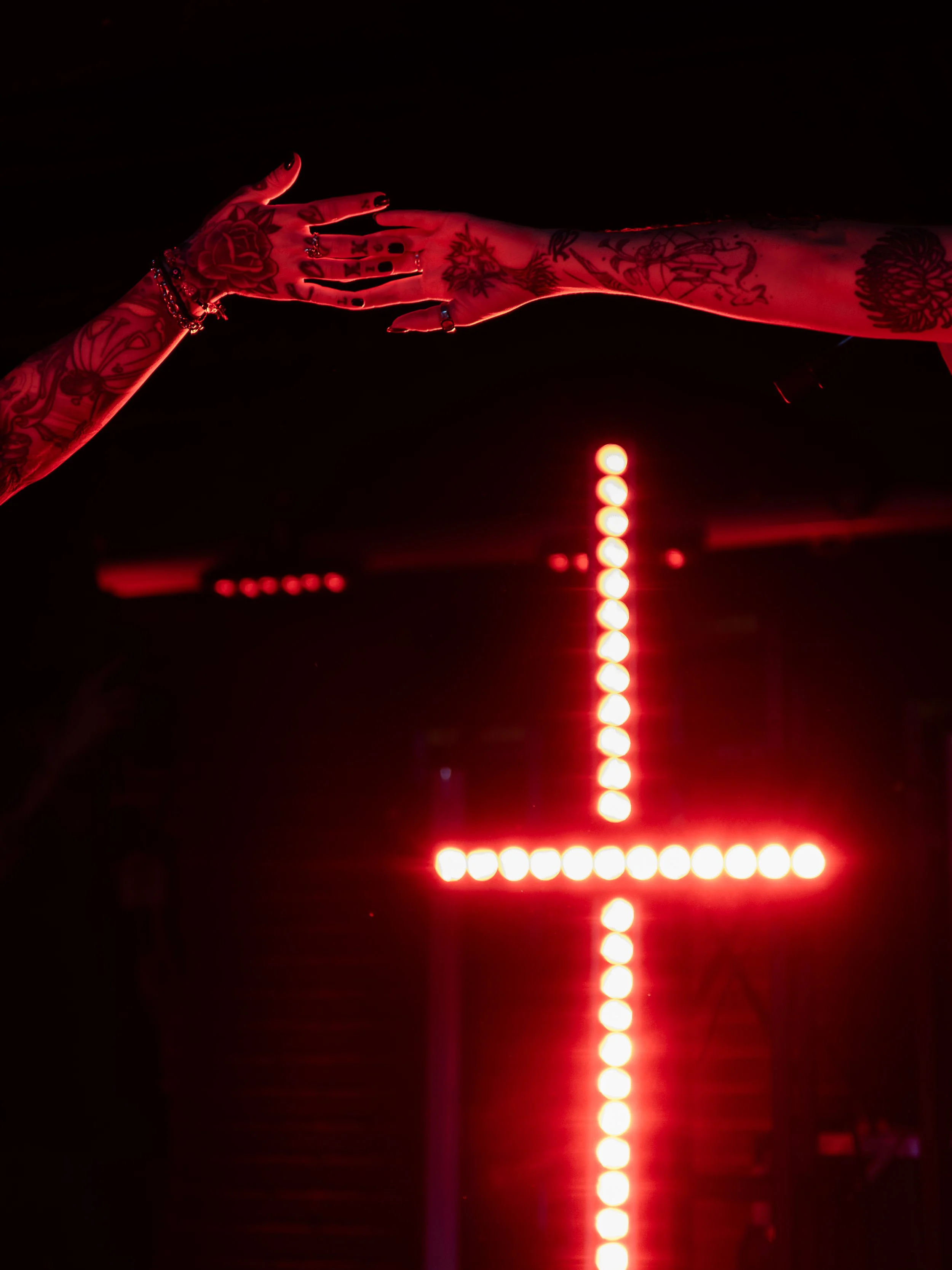 Two tattooed hands reaching out toward each other in front of a neon red cross in the dark.
