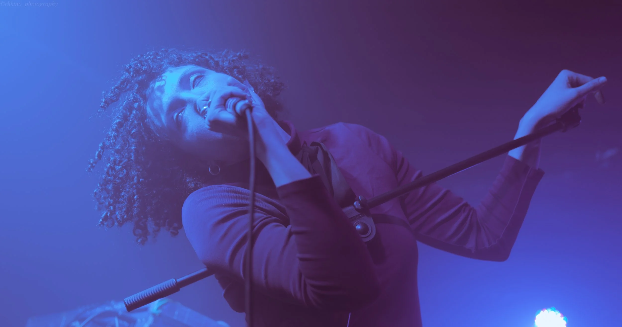 A woman with curly hair singing into a microphone on stage, holding a microphone stand, with stage lights creating purple and blue hues.
