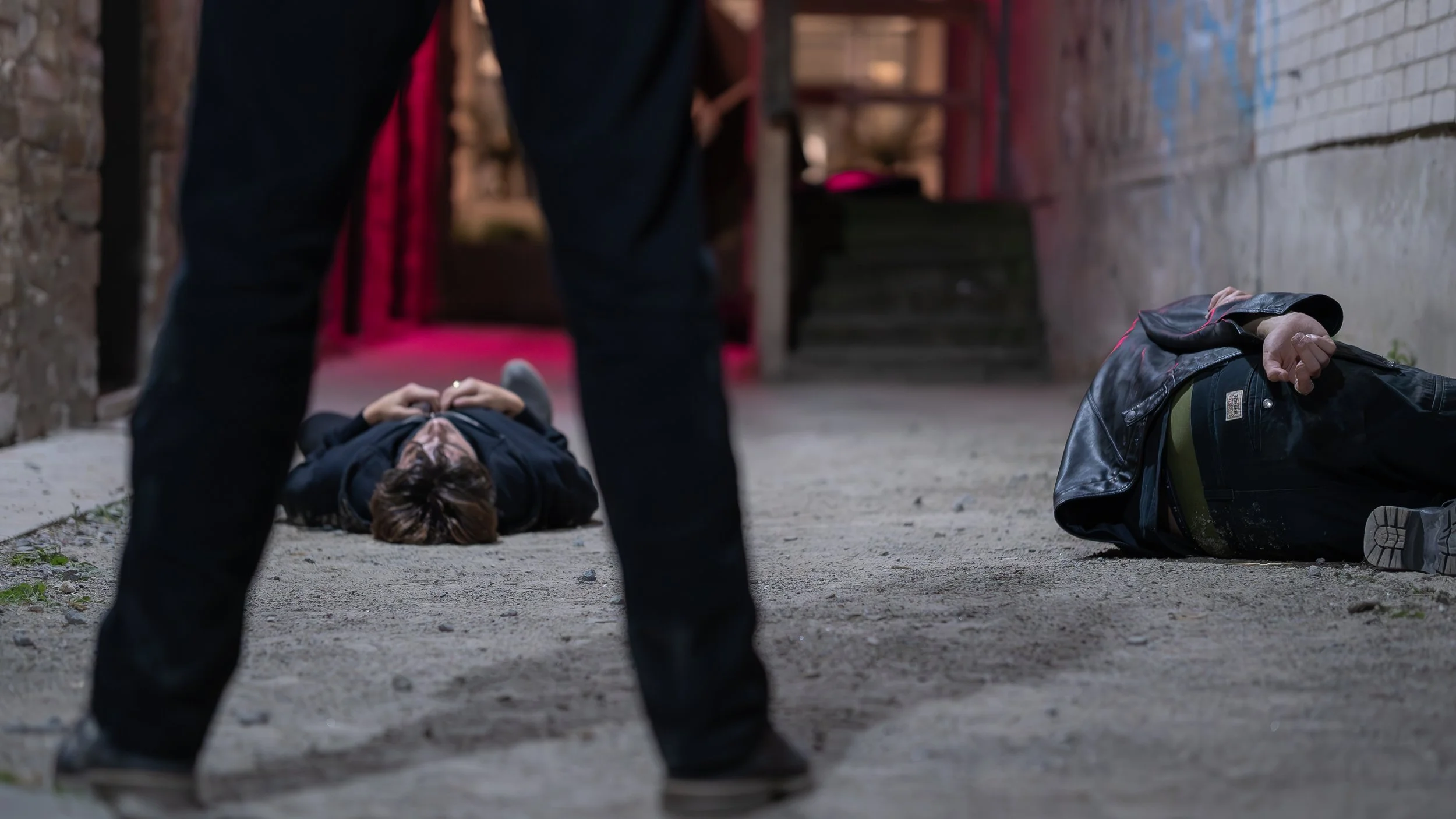 Two people are lying on the ground, face down, in an alleyway at night. A person stands between them, partially visible, with legs apart. The alley has brick walls and a dirt floor.