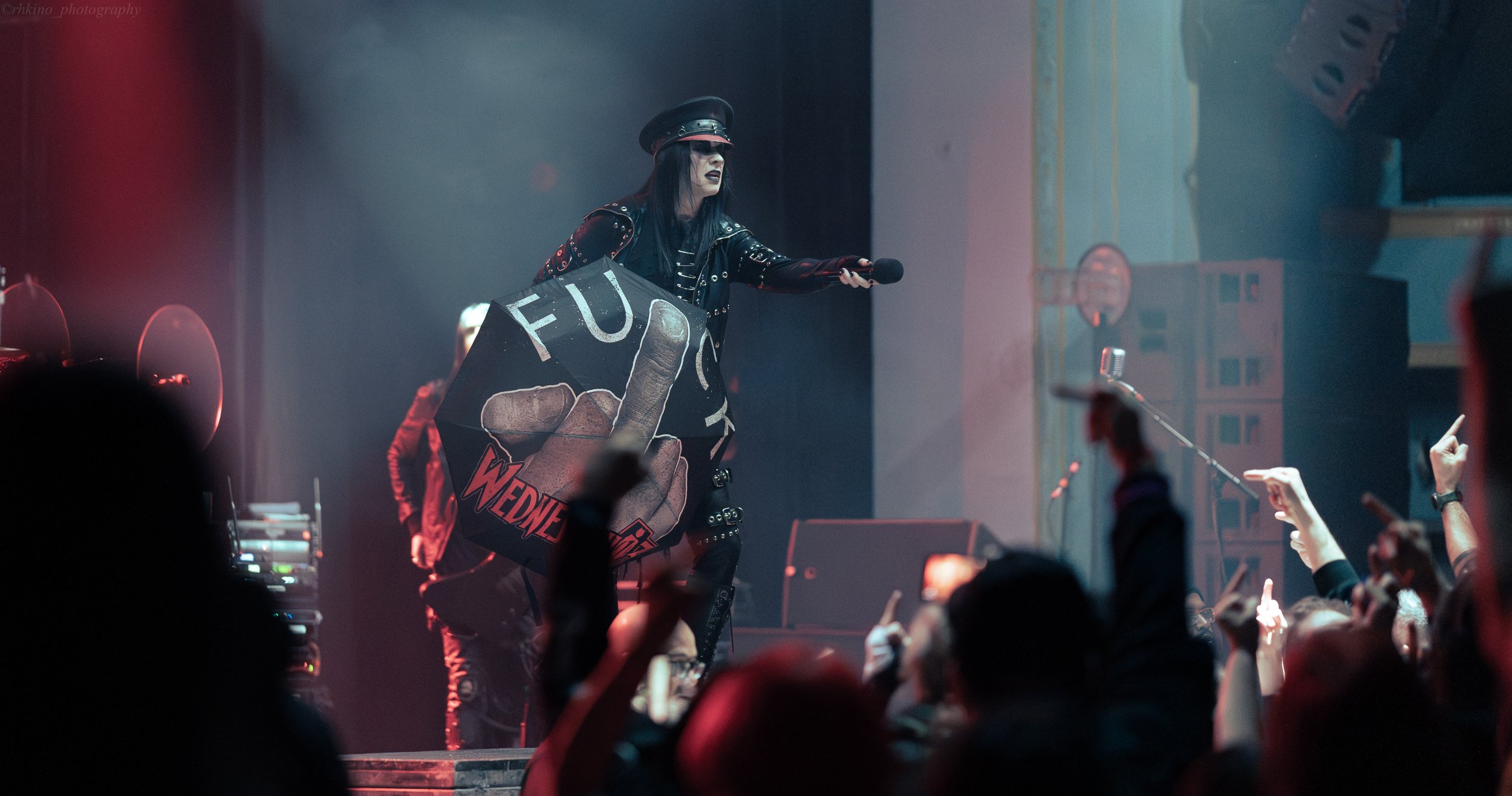 Performer on stage wearing black leather outfit and hat, holding a microphone, with a large offensive sign featuring a middle finger and the word "FUCK" in front of the audience.