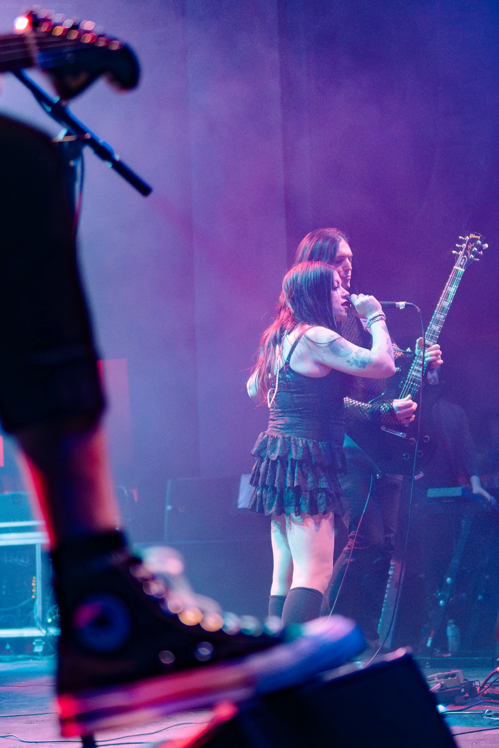 A female singer with long dark hair, wearing a black lace dress and thigh-high socks, performs on stage with two male guitarists. The scene is illuminated with colorful stage lights and a foggy atmosphere.
