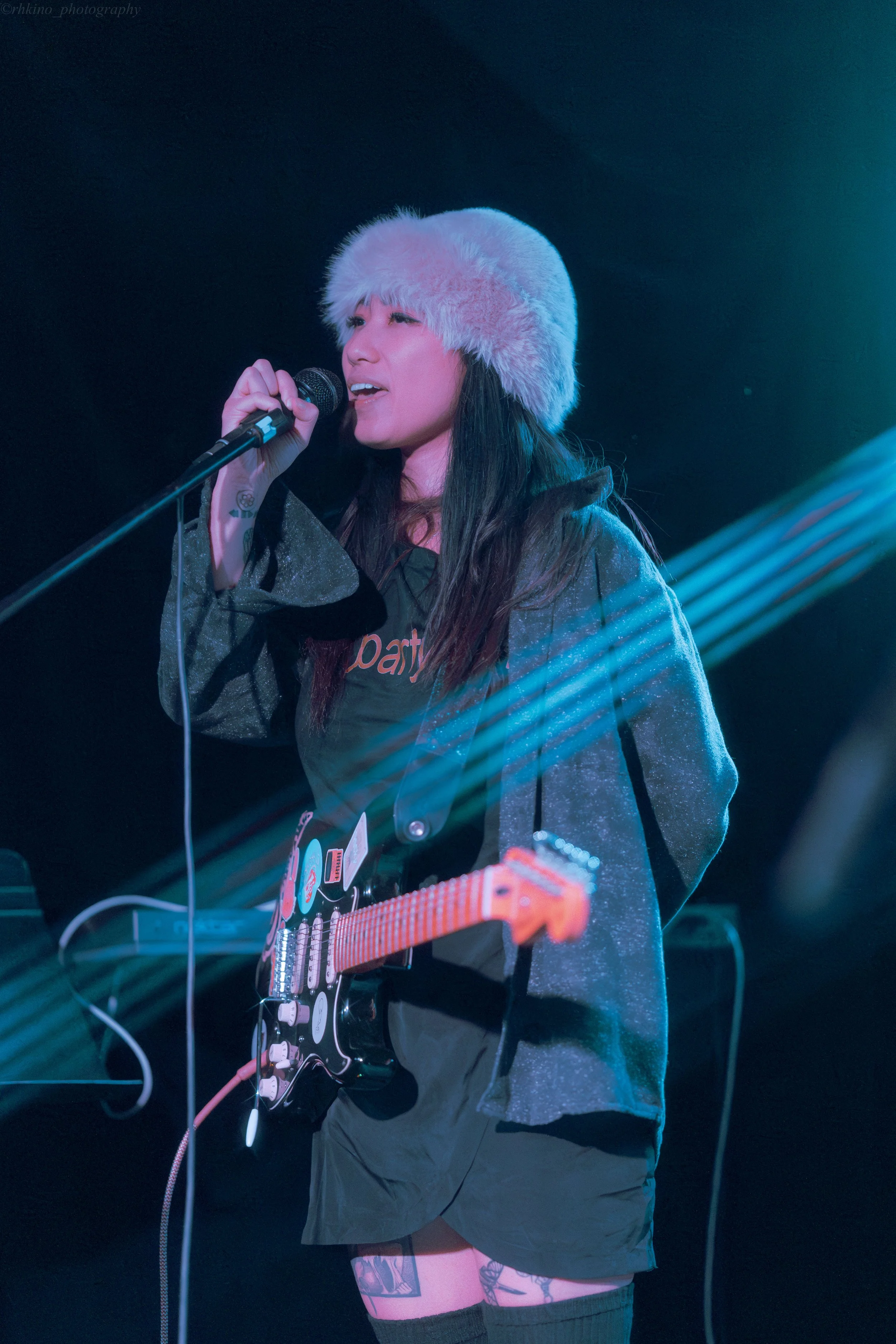 Female musician singing into a microphone while holding an electric guitar, wearing a fluffy hat and dark clothing, with stage lighting effects.