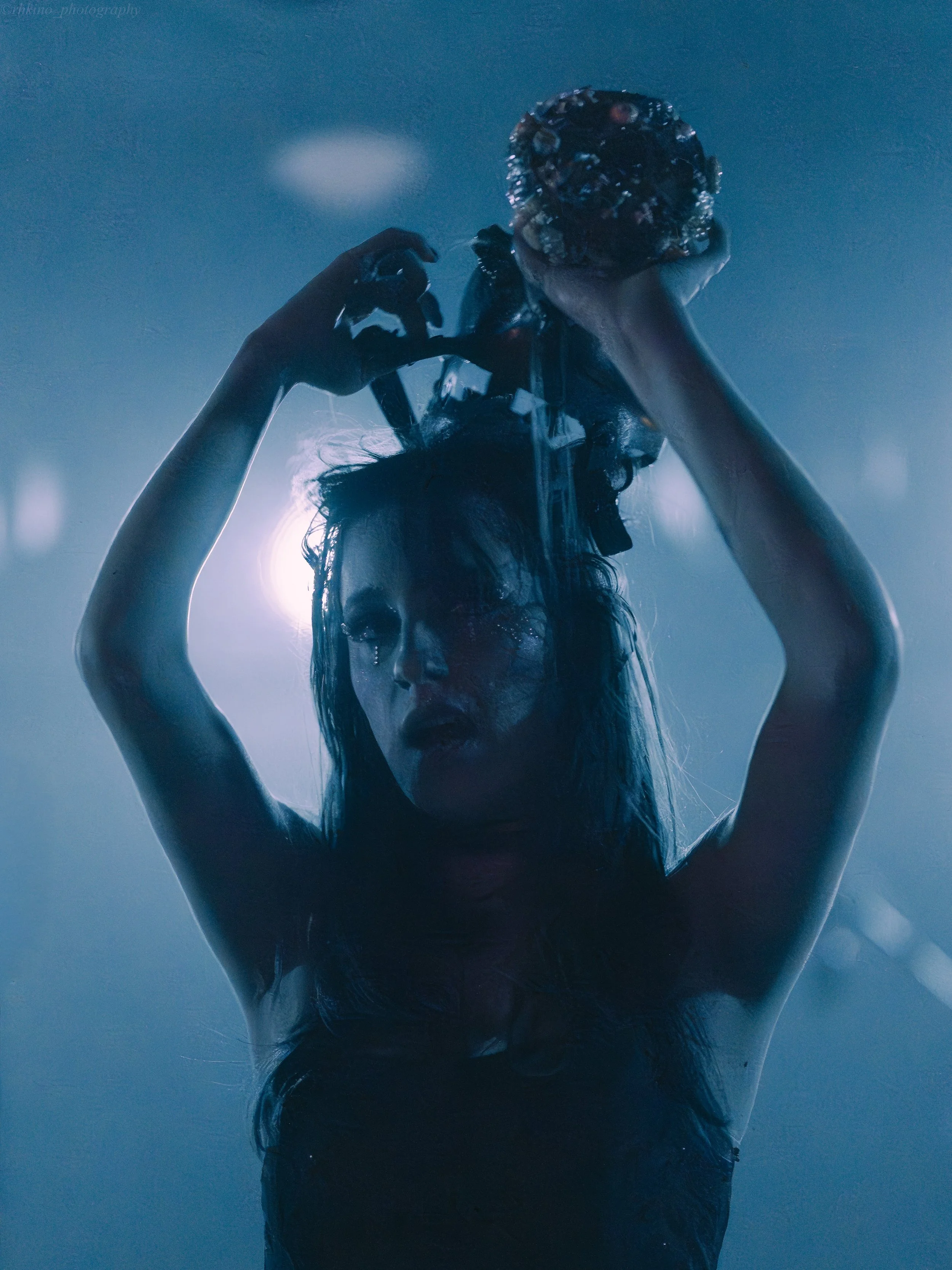 A woman with long hair and makeup holding a large apple above her head in a dark, moody setting.