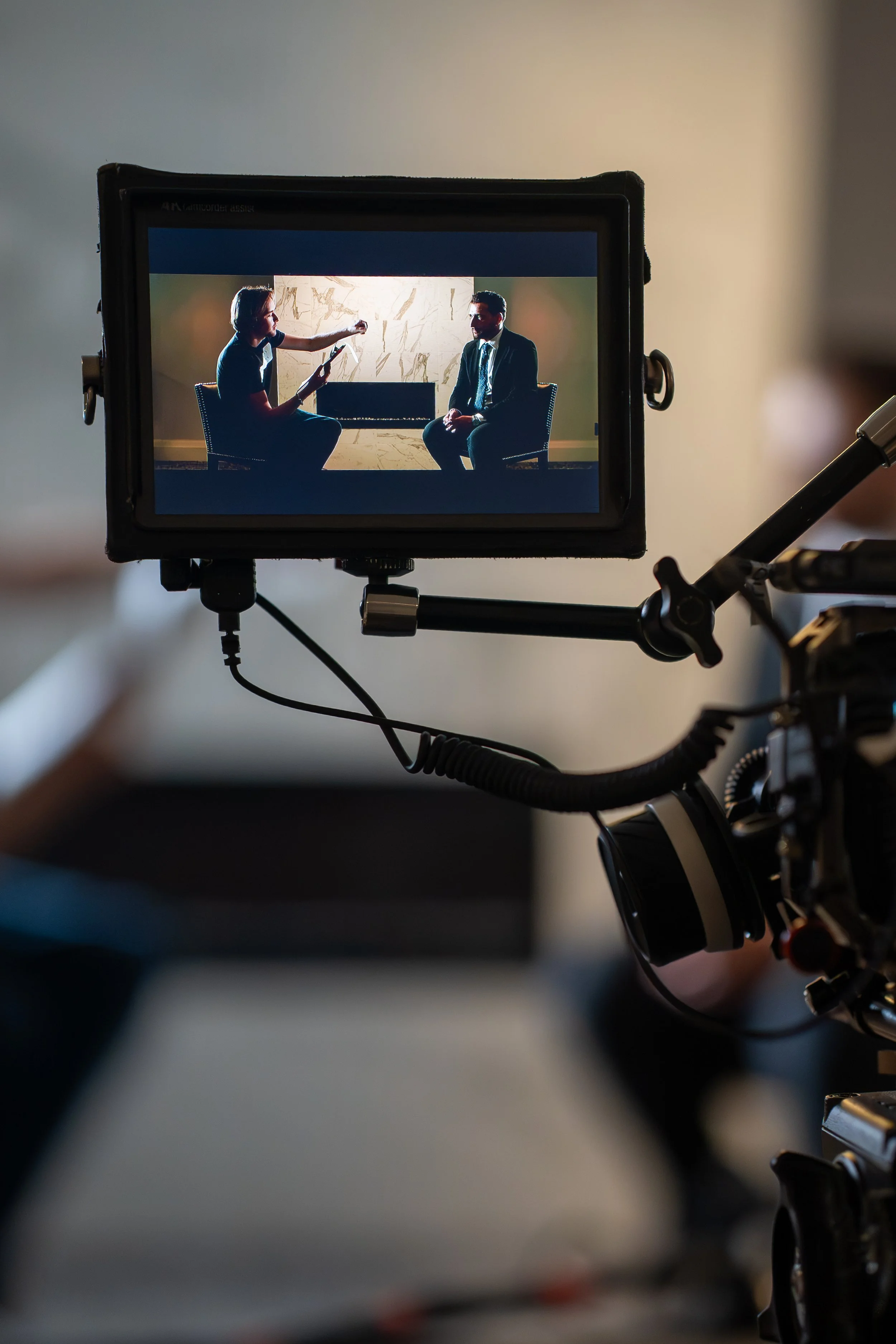 A video camera screen shows a woman interviewing a man in a professional setting, with both seated and facing each other.