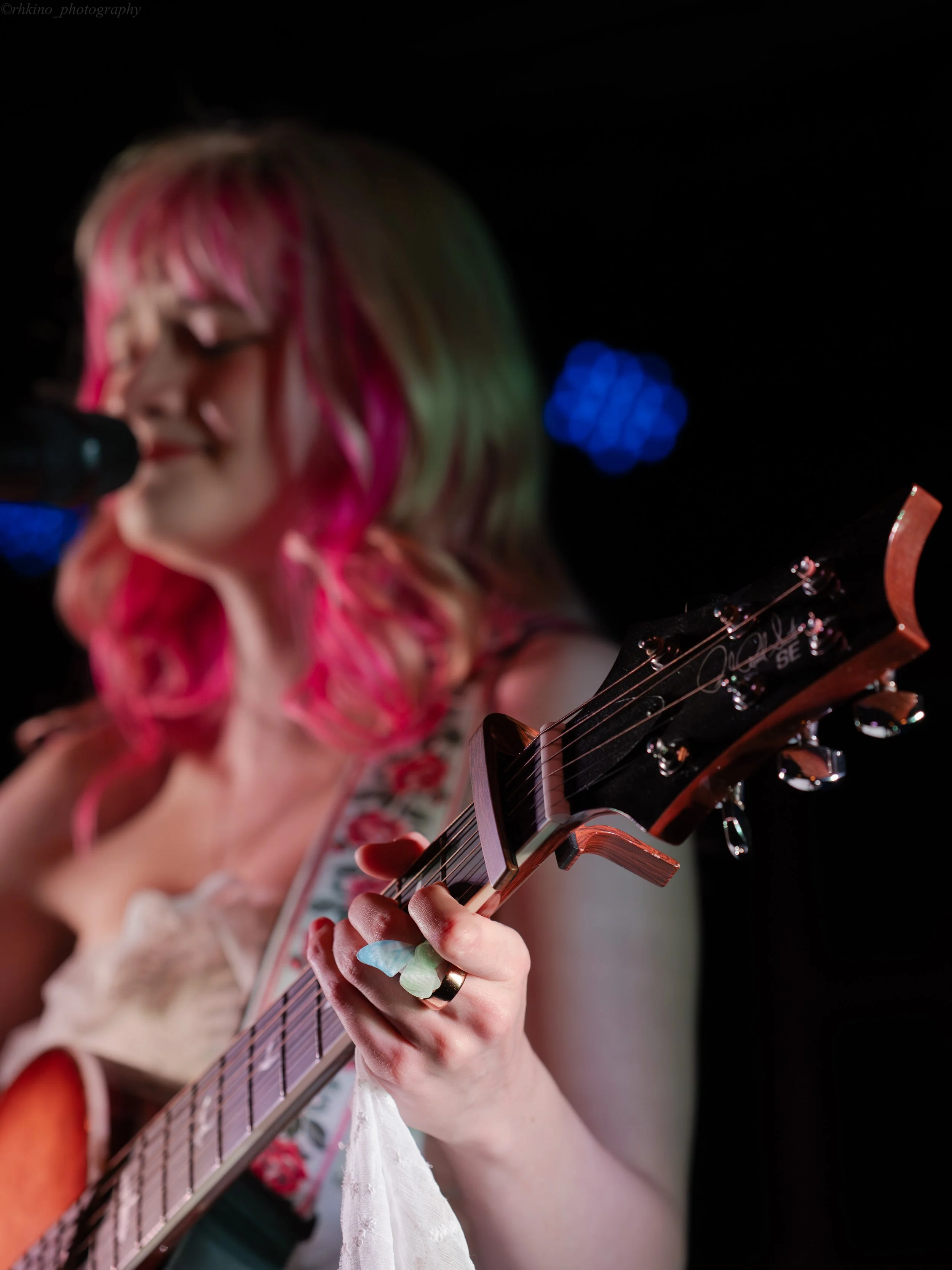 A woman with pink and green hair playing an acoustic guitar and singing into a microphone on stage, with a dark background and blue stage lights.