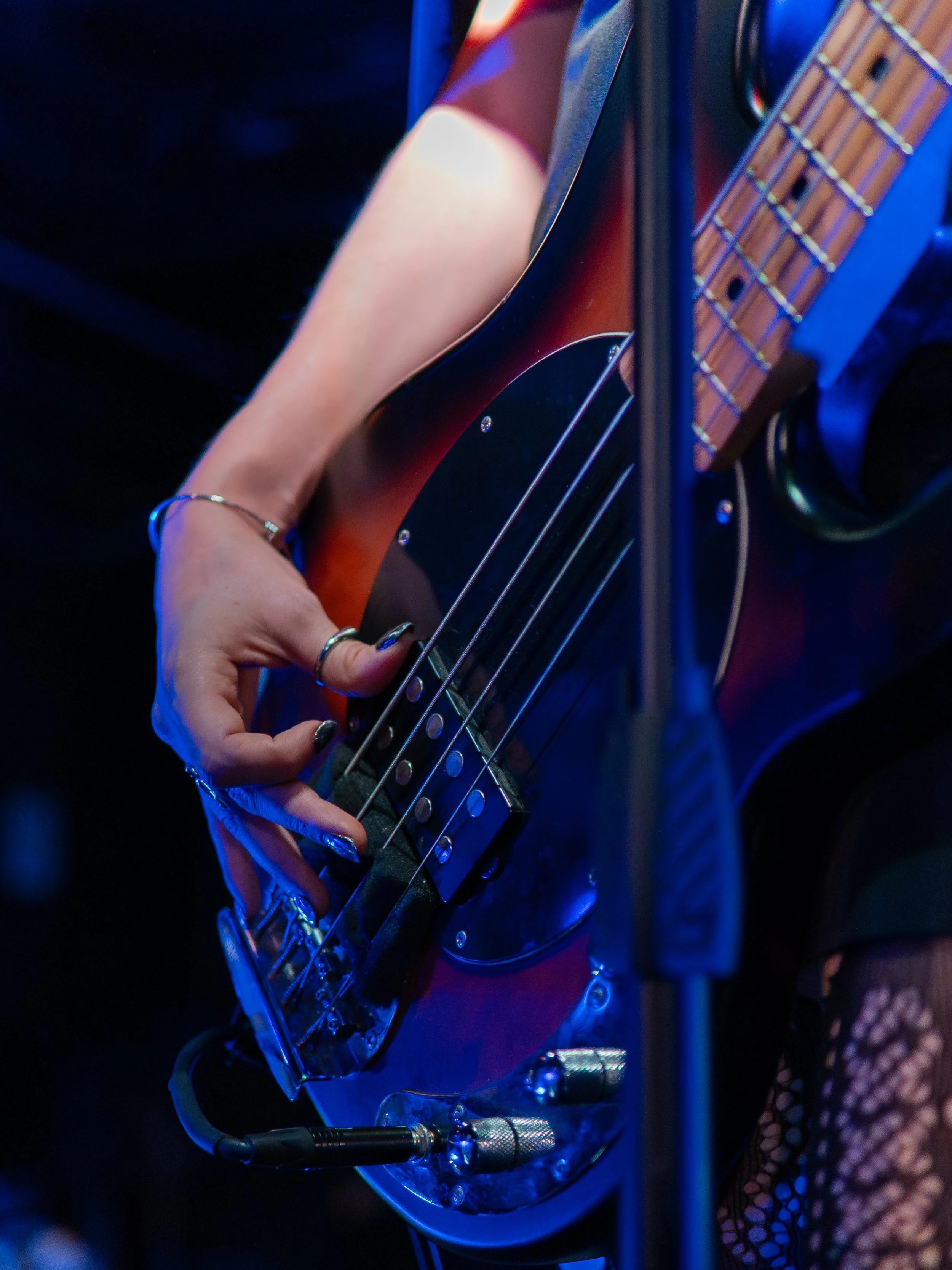 Close-up of a person playing a bass guitar on stage, with a focus on their hand on the strings and part of their arm visible.