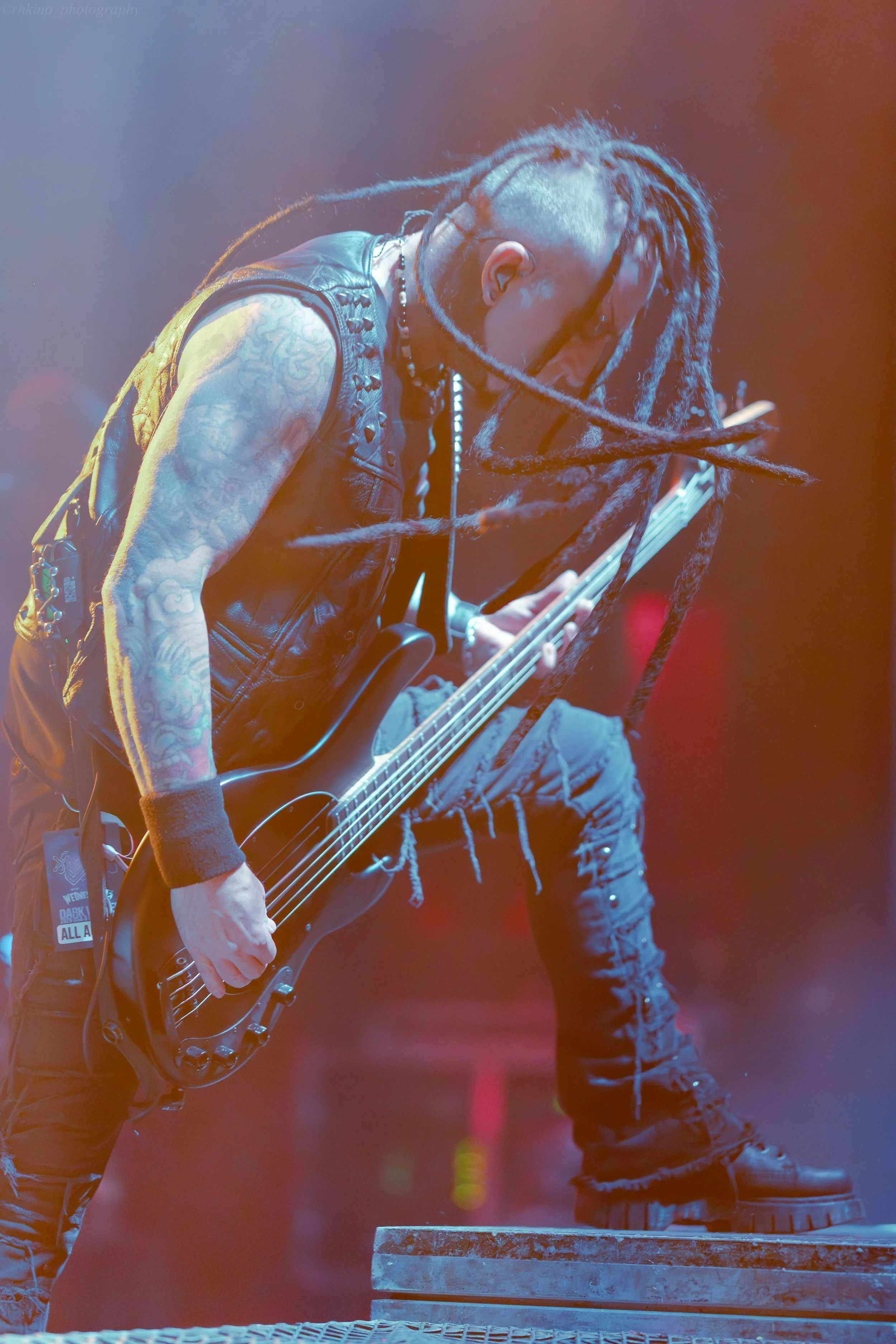 A musician with long dreadlocks, tattoos, and sunglasses playing a bass guitar on stage, wearing a black sleeveless vest, cargo pants, and black boots.
