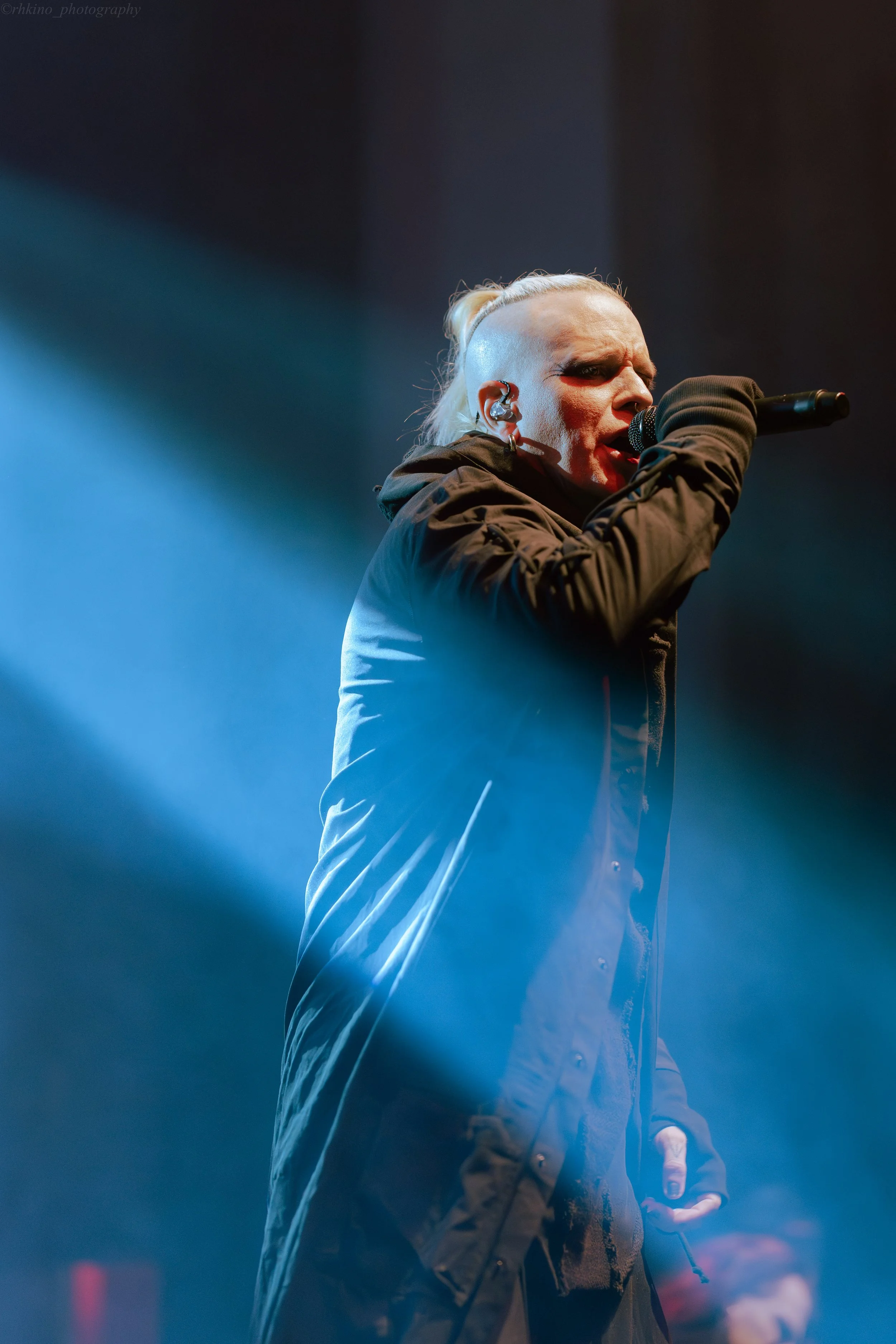 A singer with platinum blonde hair styled in a high ponytail, wearing black clothing, performs on stage with a microphone, illuminated by stage lighting and fog.