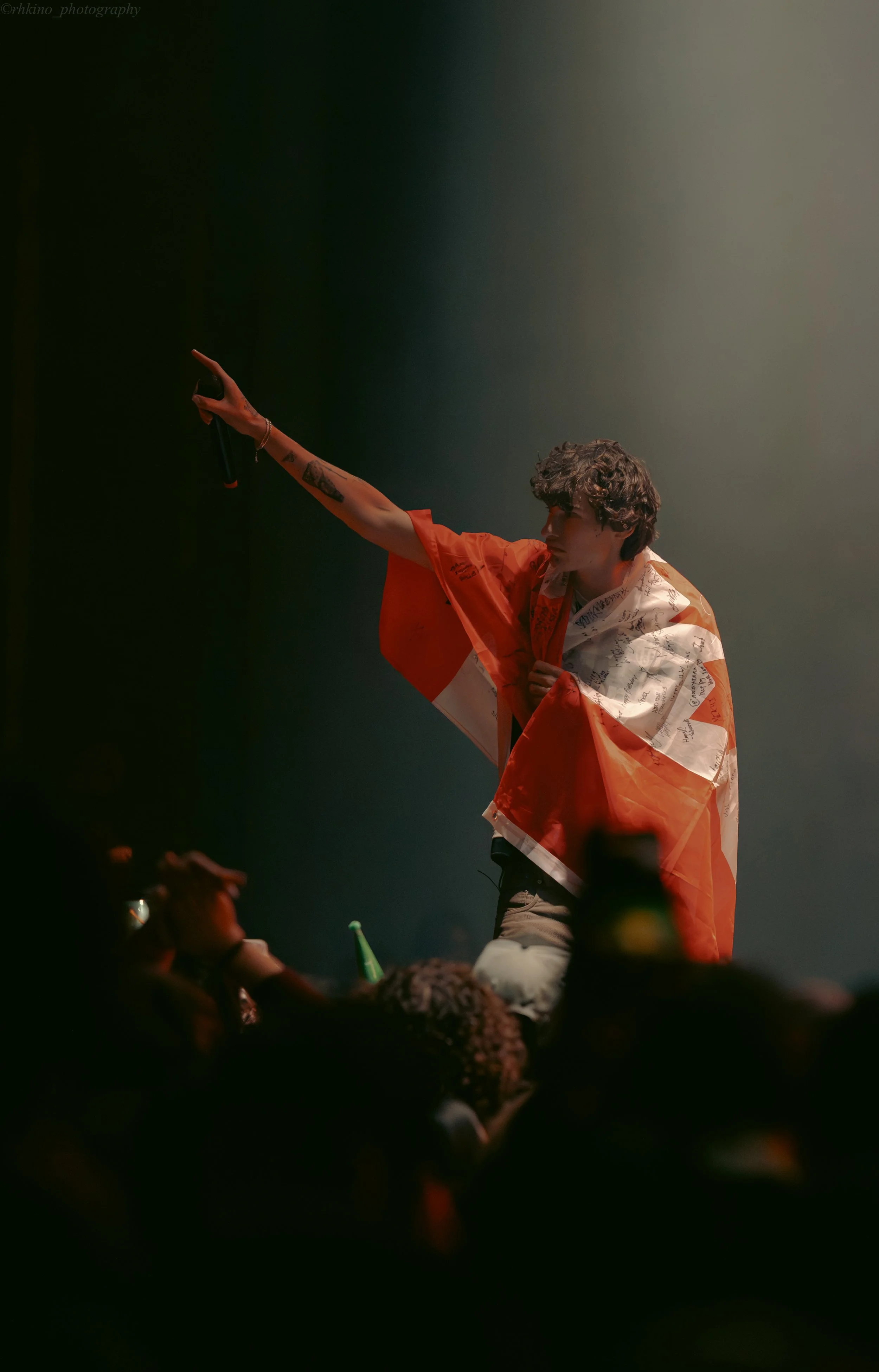 A young man with curly hair performing on stage, draped in an orange and white flag, with a microphone in hand, reaching out to the audience amid dim, smoky lighting.