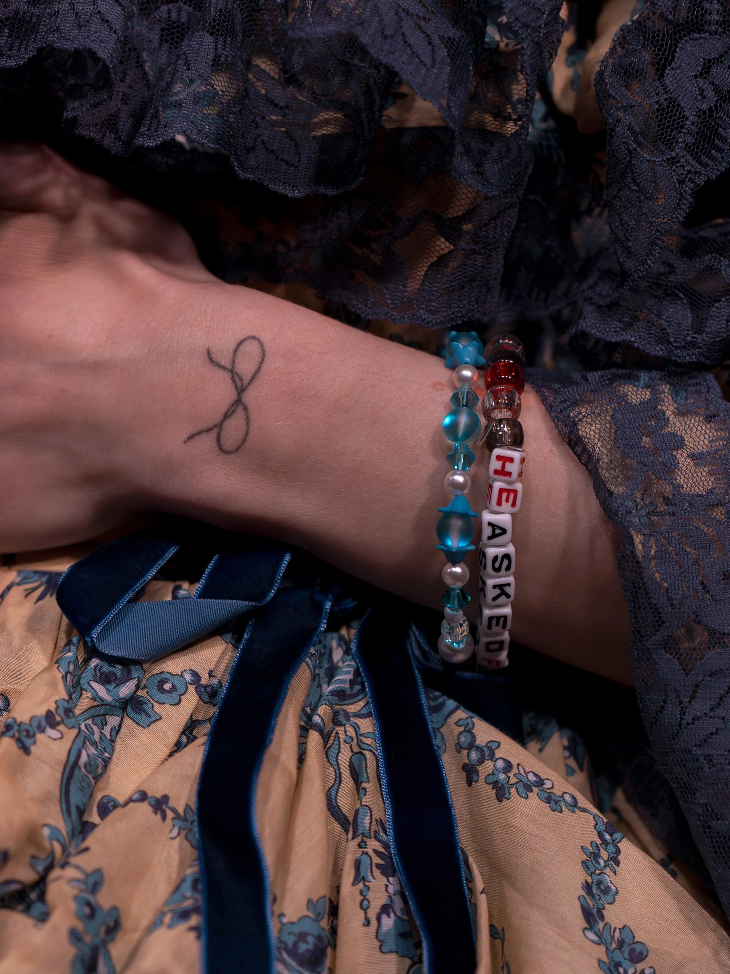 A person's wrist resting on a floral fabric with lace and a satin ribbon. The wrist has three bracelets and a tattoo of a simple stick figure.