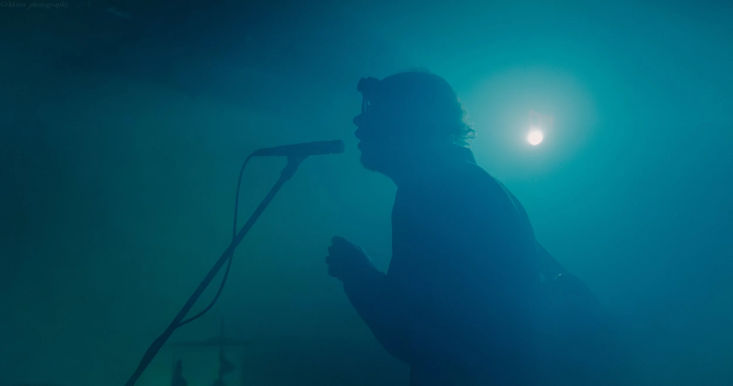 Silhouette of a person singing into a microphone on stage with a blue background and bright light behind them.