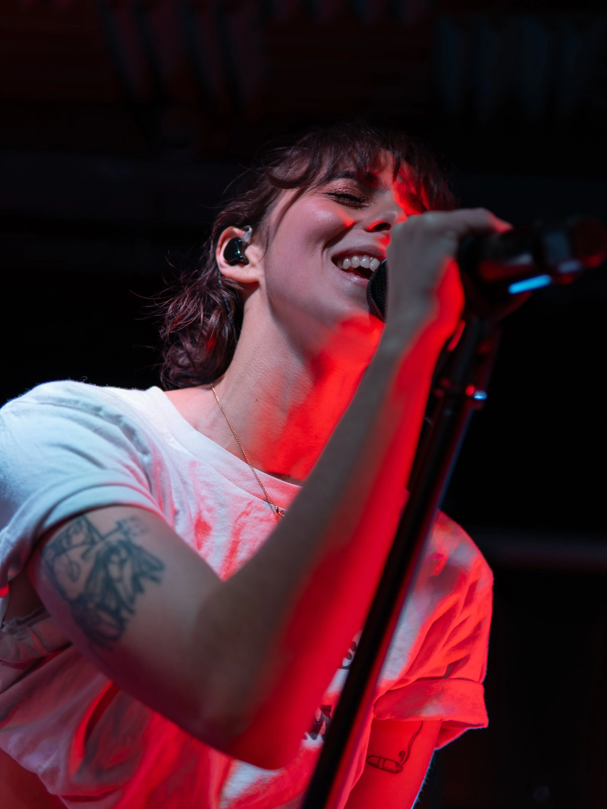 A young woman singing into a microphone on stage, smiling with eyes closed, wearing a white t-shirt, with a tattoo on her left arm, and a necklace, illuminated by stage red lighting.