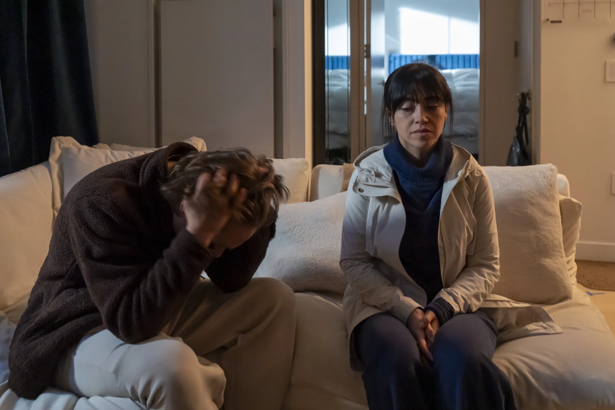Two women sitting on a couch in a dimly lit room; one woman on the left is holding her head in her hands, looking distressed, while the woman on the right, dressed in a white jacket and blue sweater, looks somber and reflective.
