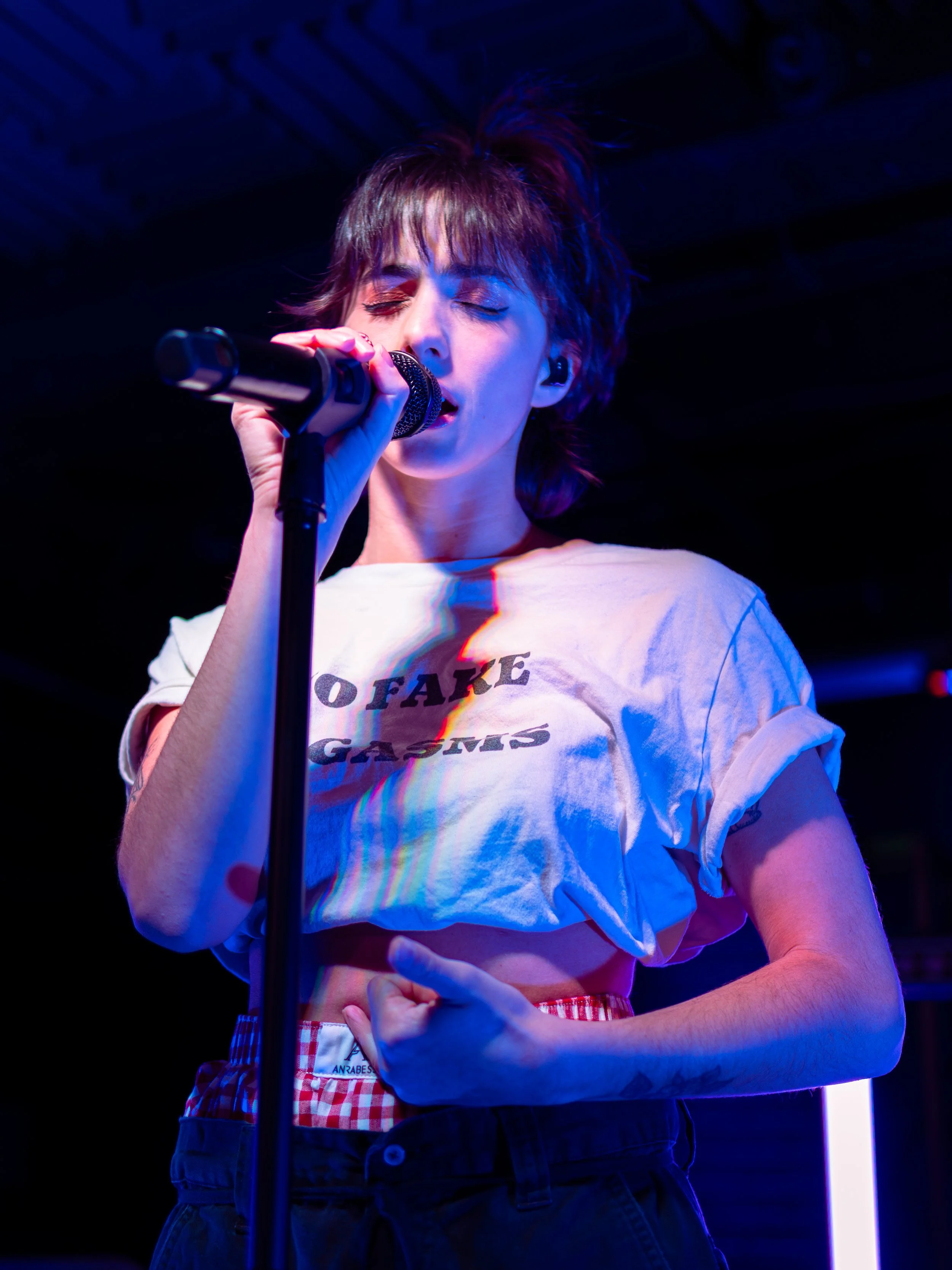 A woman singing into a microphone on stage, wearing a white t-shirt with text, and a plaid belt.