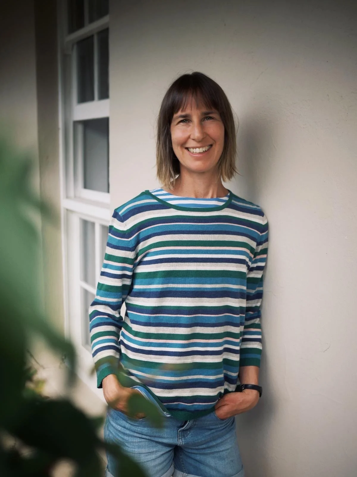 A woman smiling and standing against a light-colored wall with a window in the background, wearing a striped shirt and jeans.