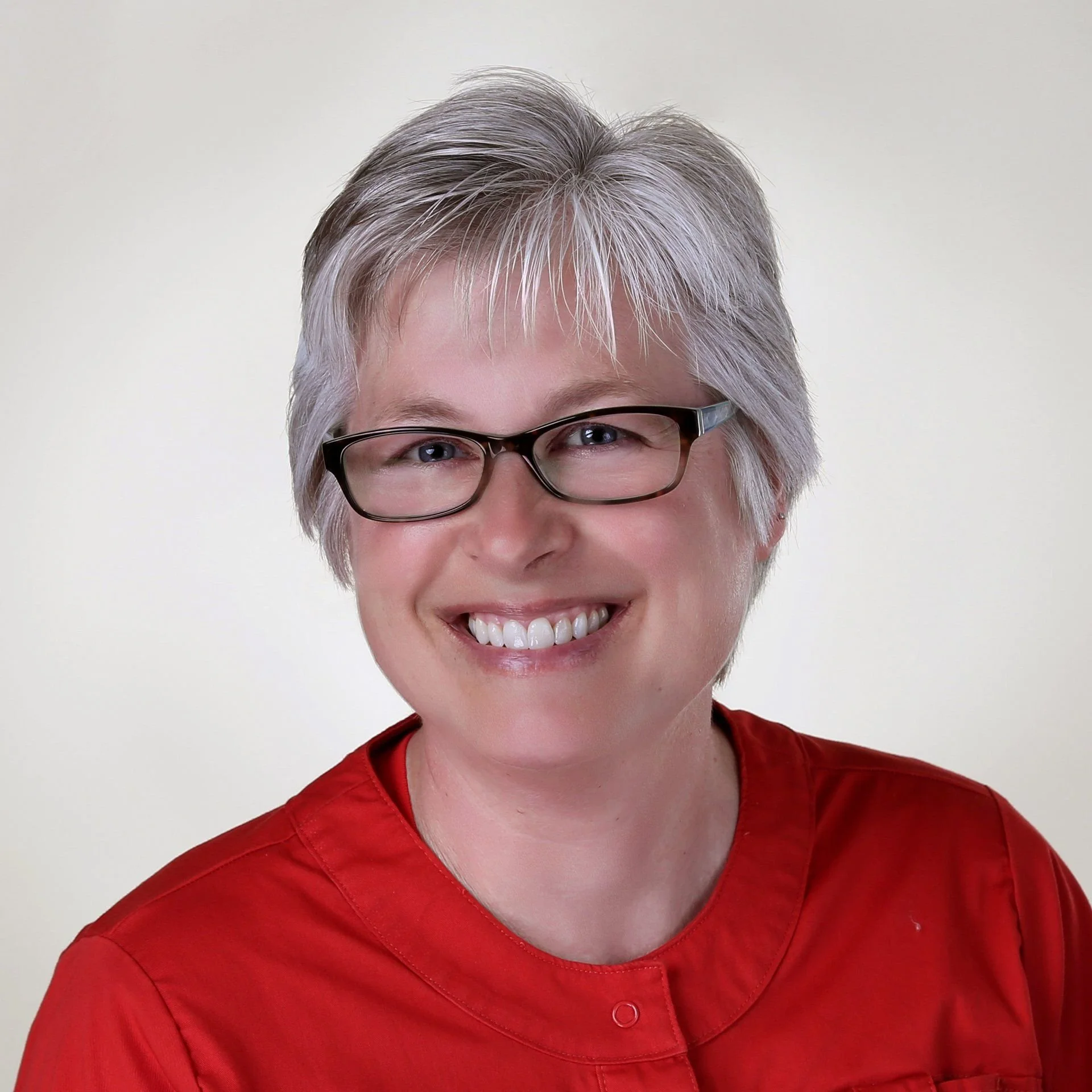 Portrait of a smiling woman with short gray hair, glasses, and a red medical uniform against a plain light background.