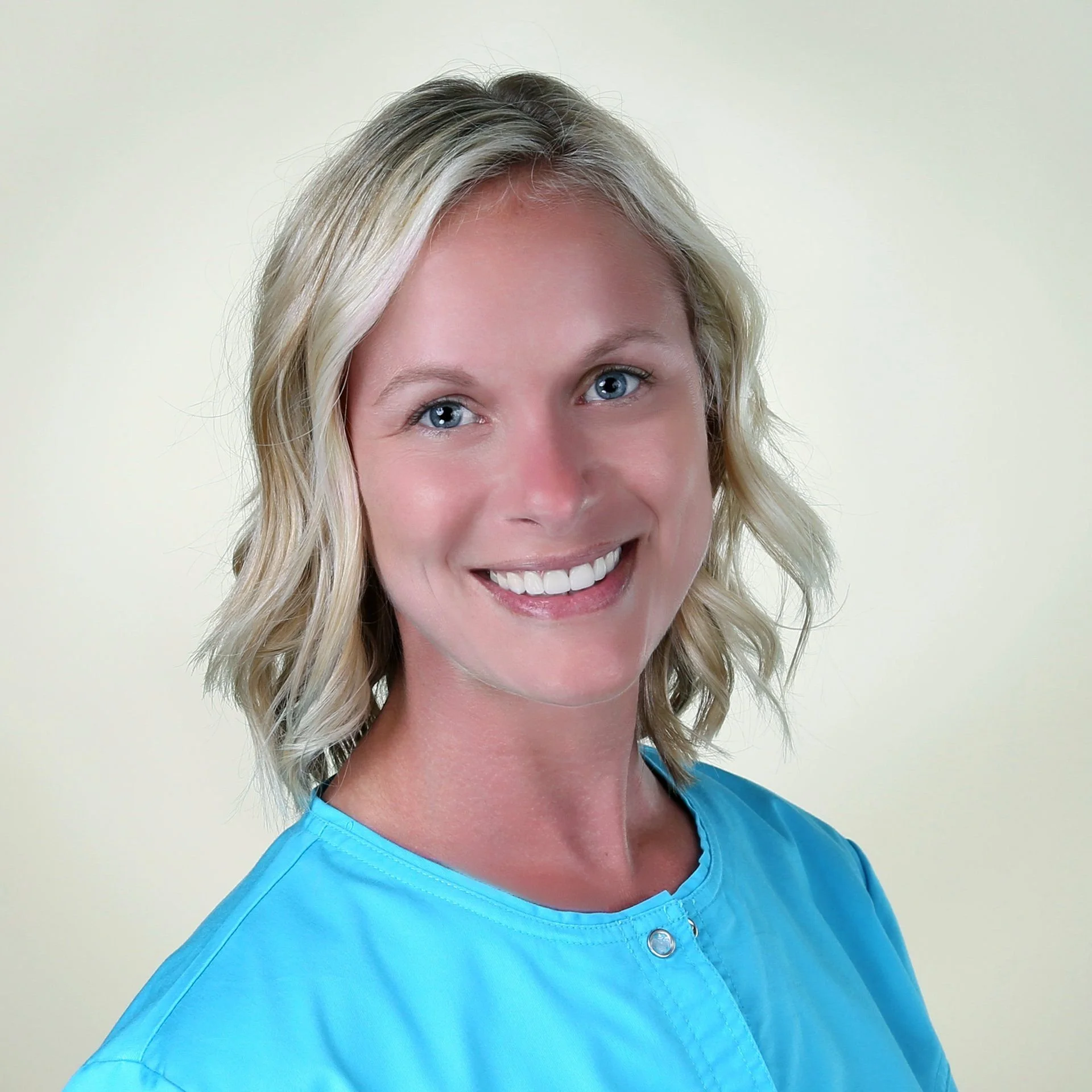 Portrait of a smiling woman with blonde hair wearing a light blue medical scrub top against a plain beige background.