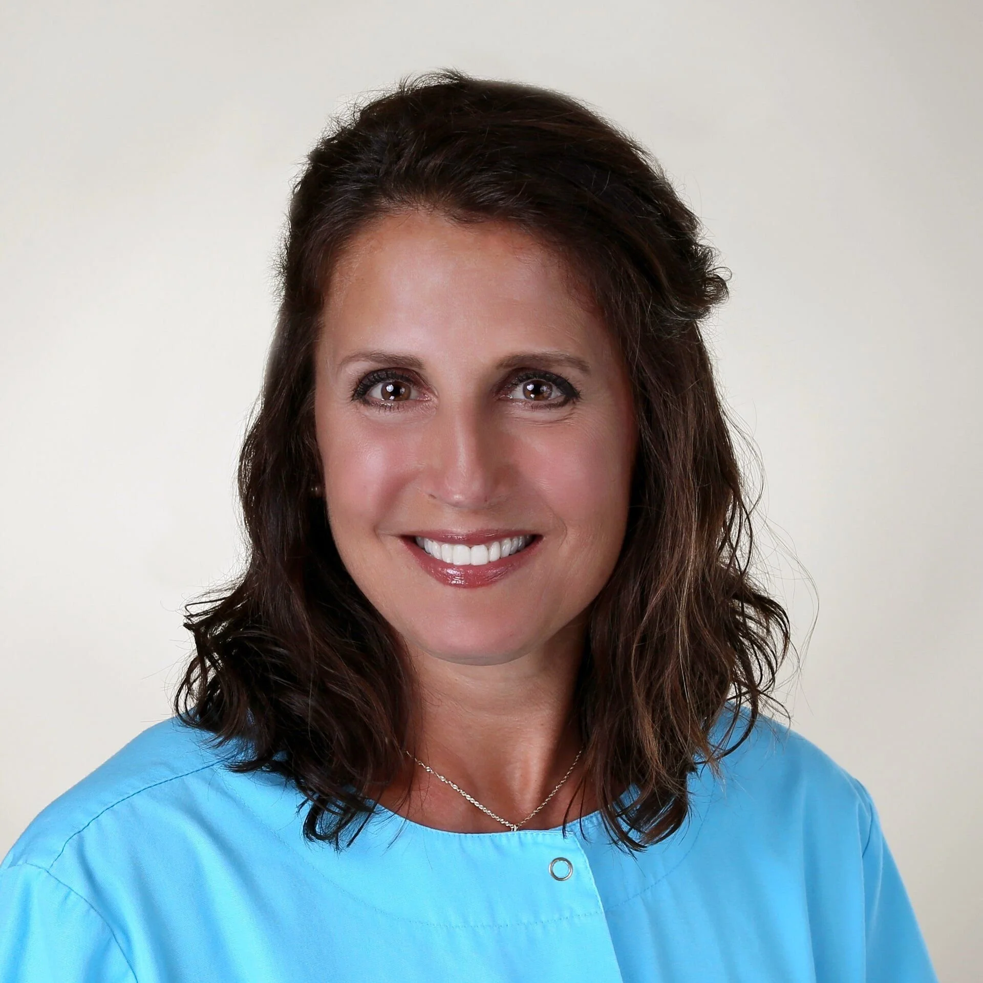 Portrait of a smiling woman with wavy brown hair, wearing a blue medical uniform and a delicate necklace, against a light background.