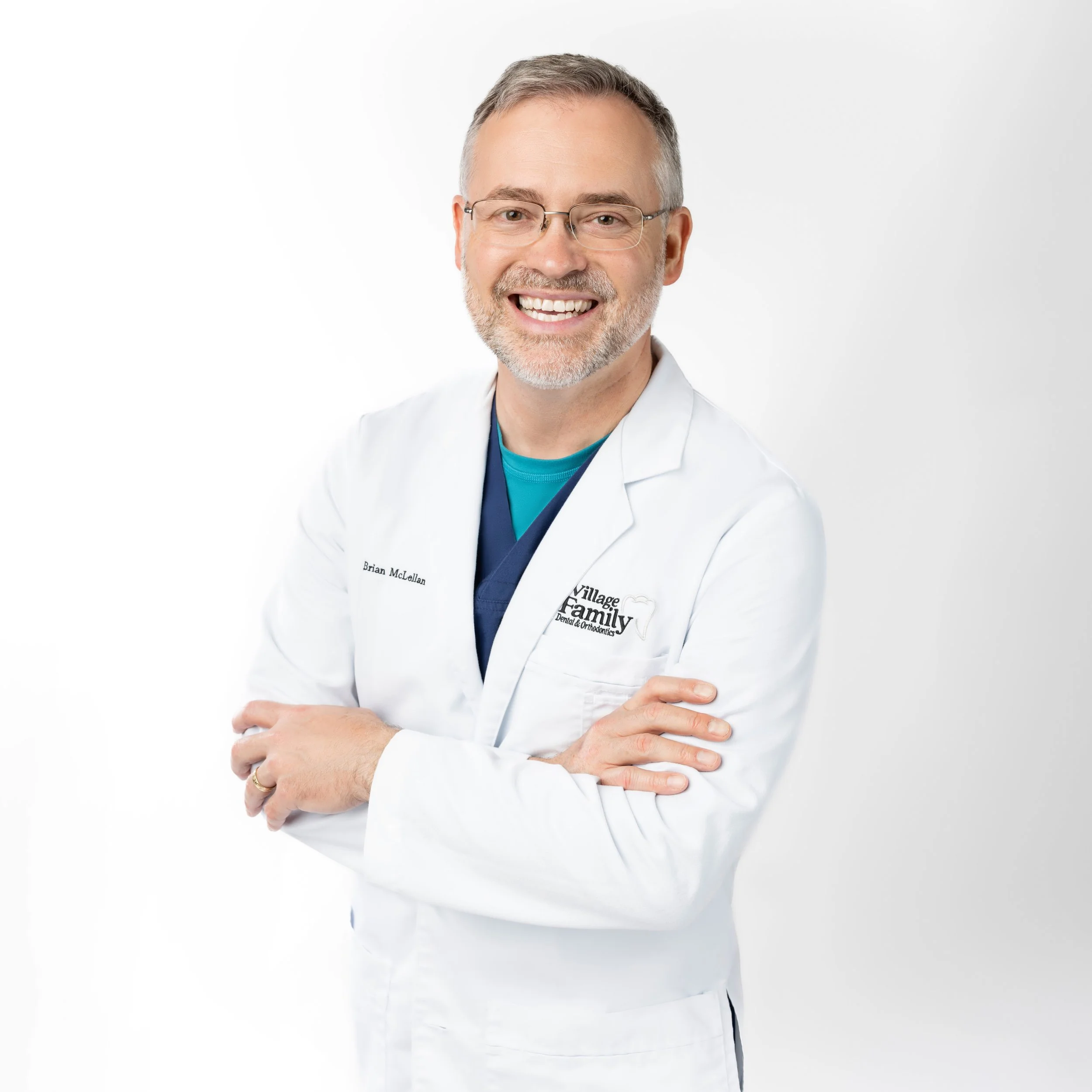 Smiling male healthcare professional with glasses and gray hair, wearing a white lab coat with "Village Family Dental & Orthodontics" embroidered on it, standing against a plain white background with arms crossed.