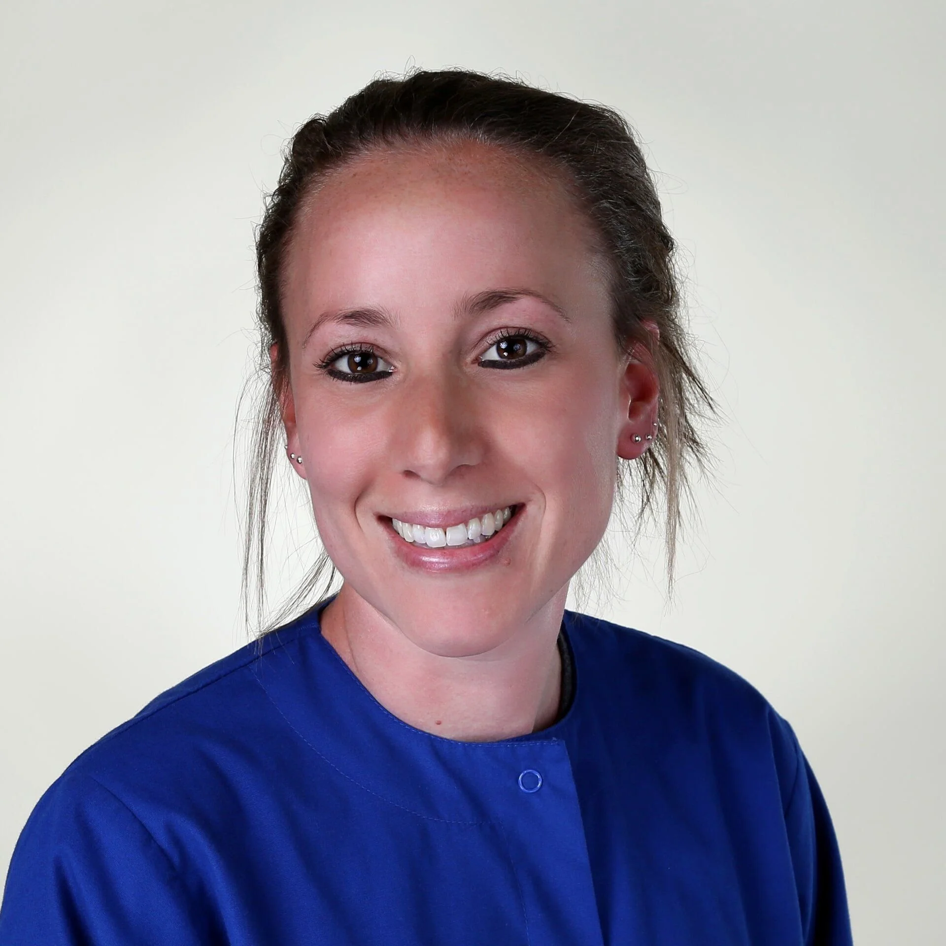 Smiling woman with light skin, brown eyes, and brown hair tied back, wearing blue medical scrubs against a plain background.
