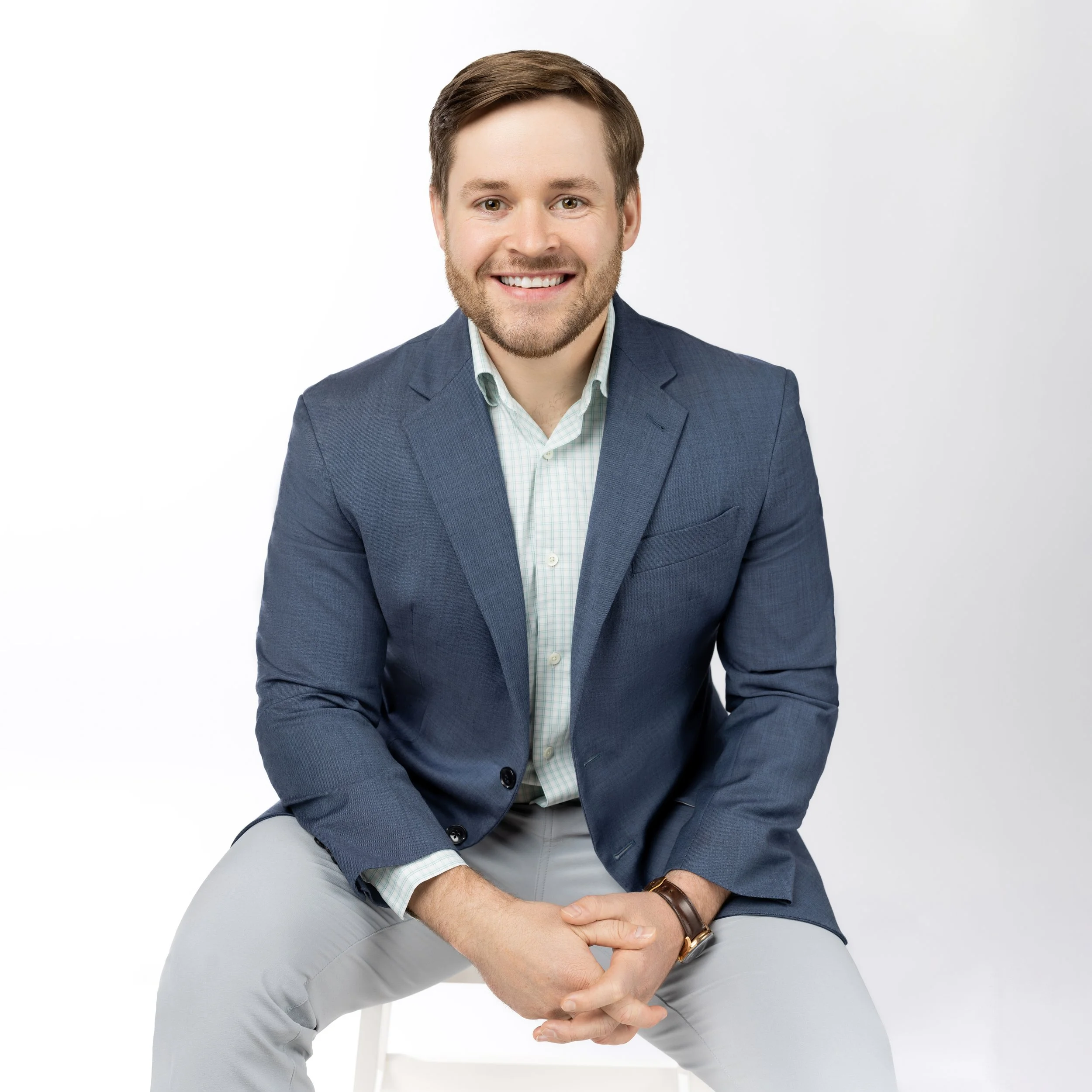 A smiling man with light brown hair and a beard, wearing a blue blazer, light green checkered shirt, and light gray pants, sitting against a white background.
