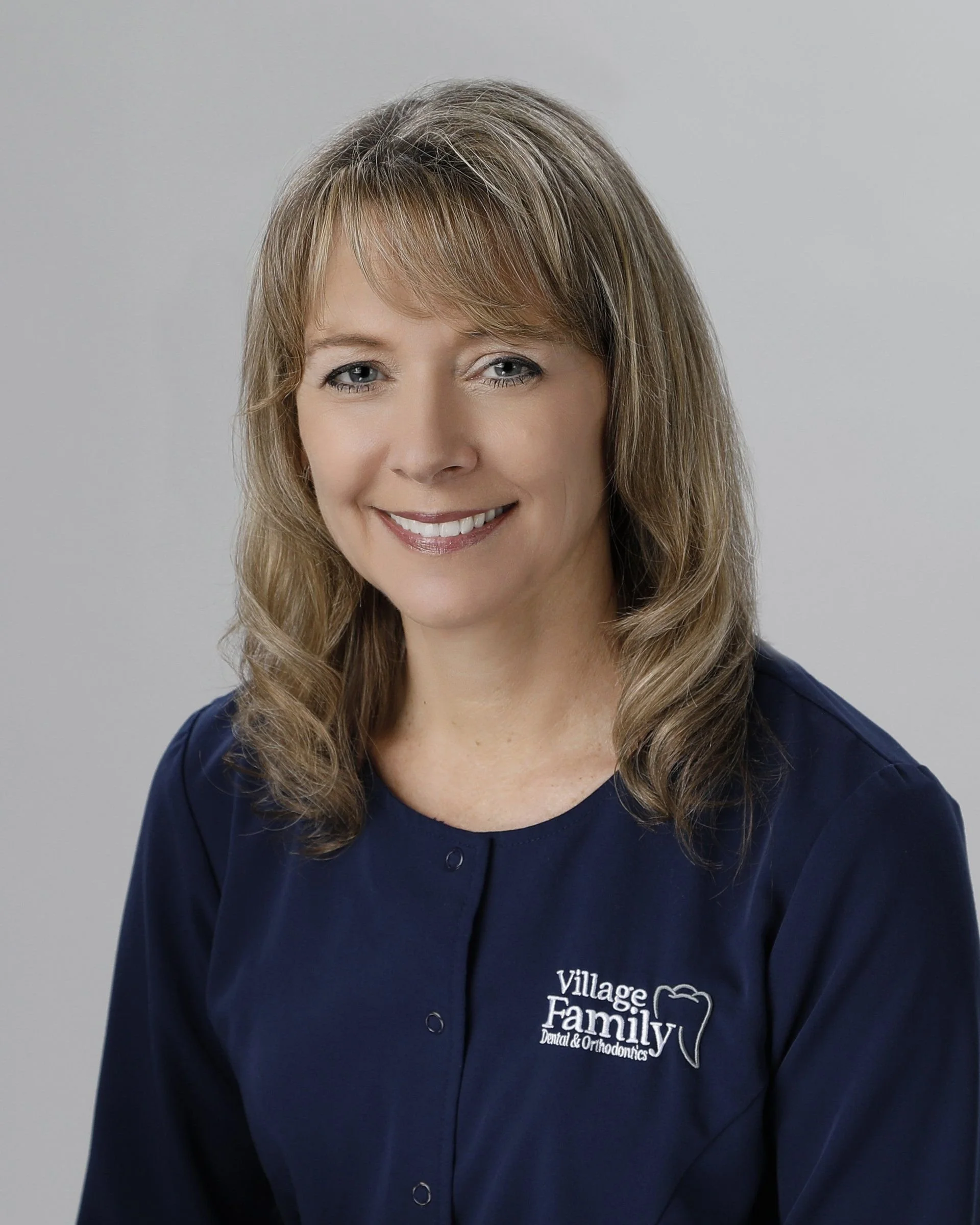 A woman with light brown, wavy hair smiling at the camera wearing a navy blue jacket with a Village Family Dental & Orthodontics logo.