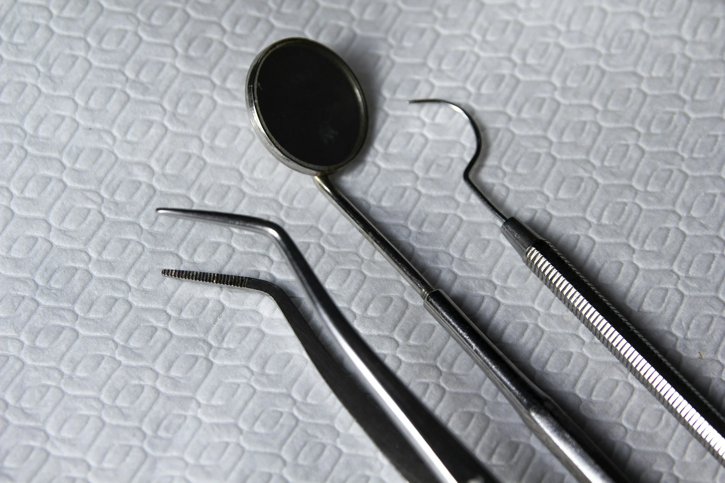 A dental mirror, a dental probe, and tweezers are placed on a textured white surface.