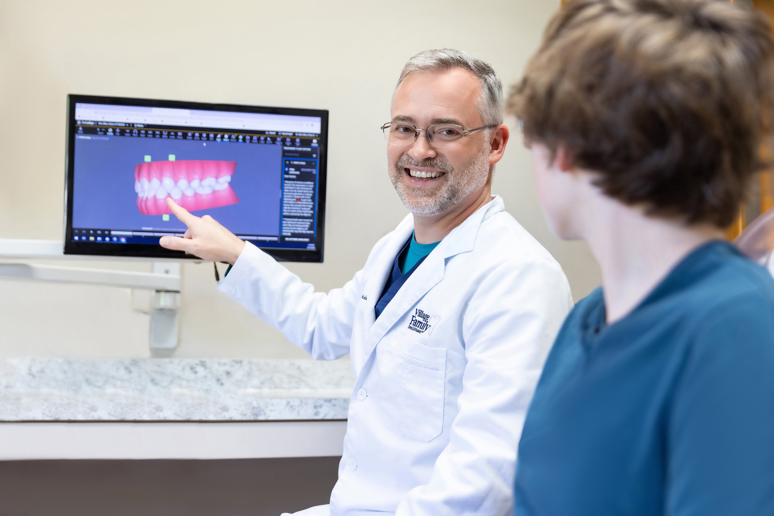 Dentist in white coat pointing at a computer screen displaying a 3D model of teeth to a patient in a dental office.
